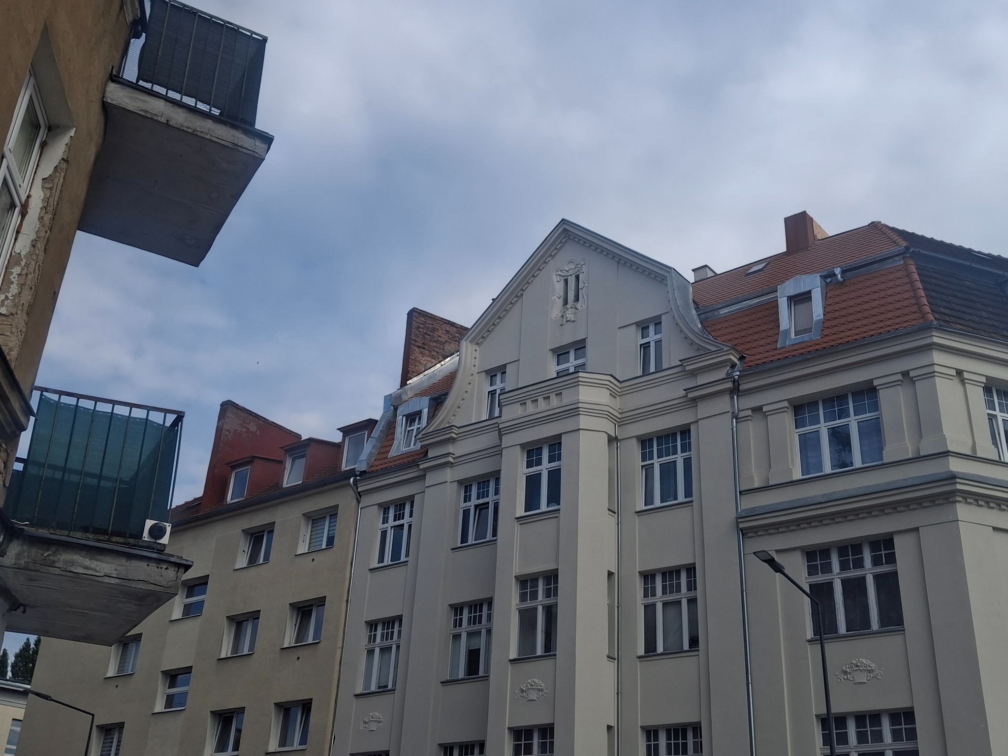 Elewacja kamienicy z czerwonym dachem i oknami dachowymi, widziana z dołu. Na pierwszym planie fragment balkonu sąsiedniego budynku.