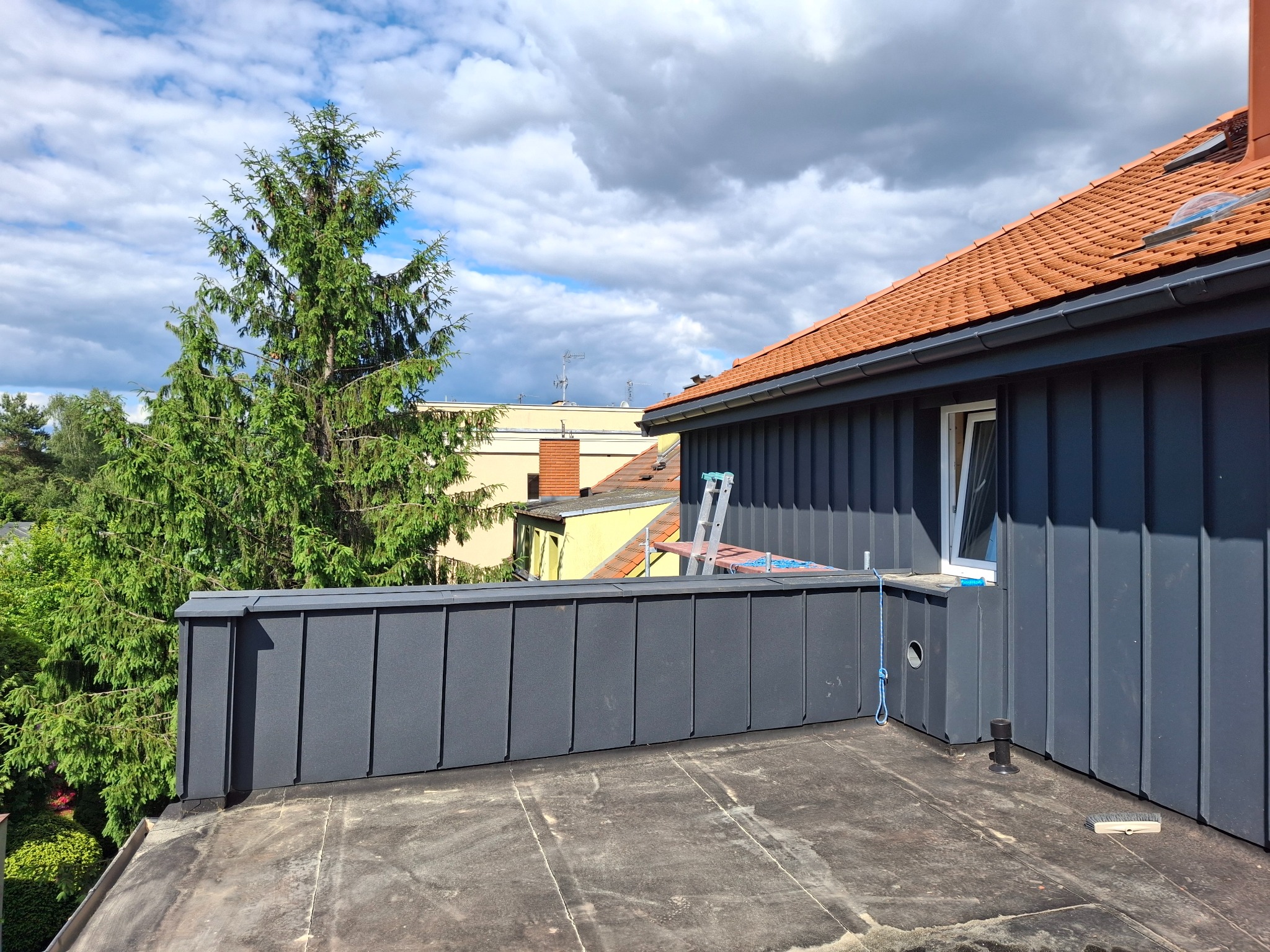 Szary dach z blachy na tle zieleni i błękitnego nieba z chmurami. Widoczna drabina i fragment dachu z czerwoną dachówką.