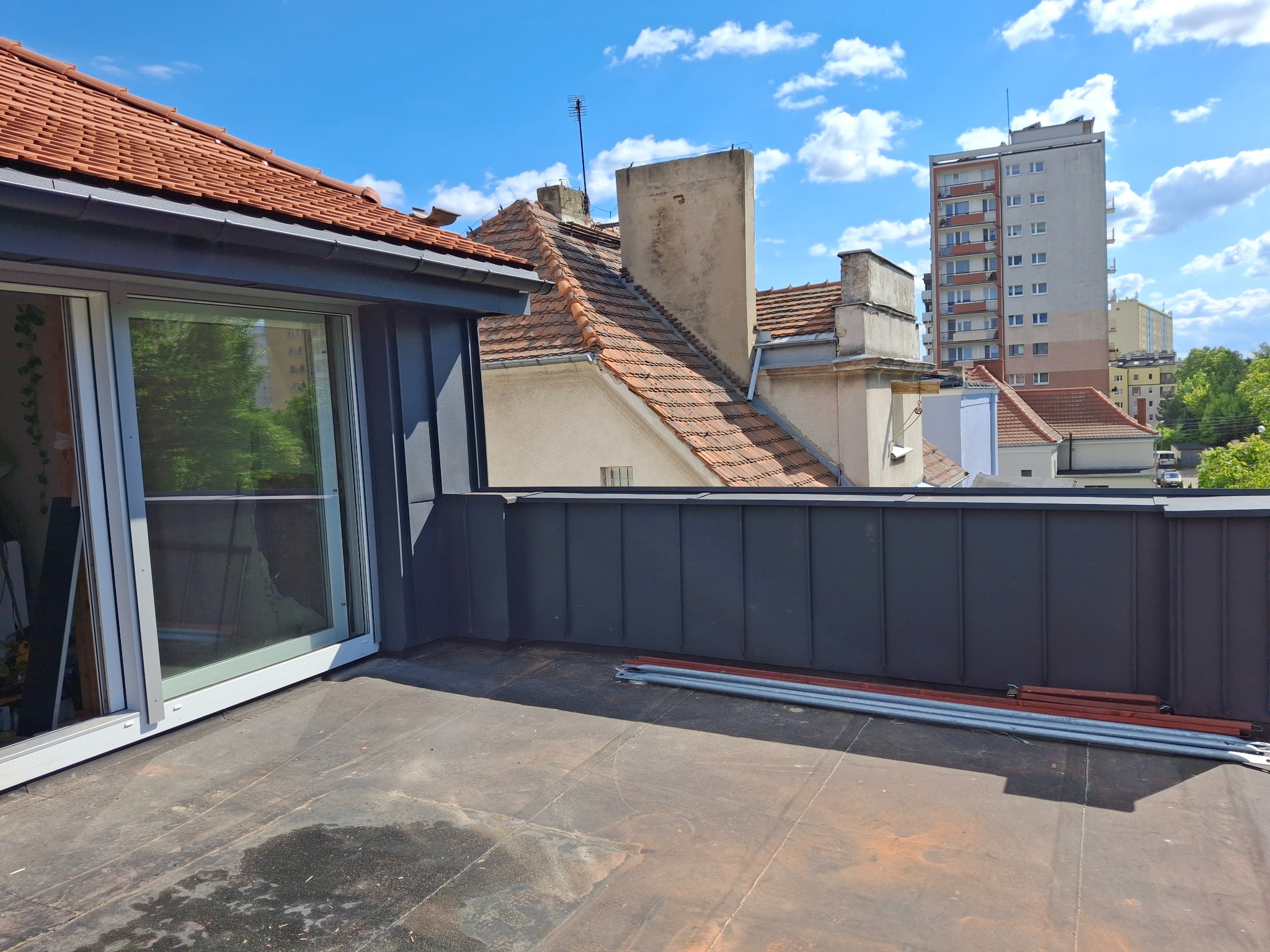 Dach pokryty papą z obróbką blacharską w kolorze grafitowym, widok na budynki w tle i błękitne niebo. Na dachu leżą rury.