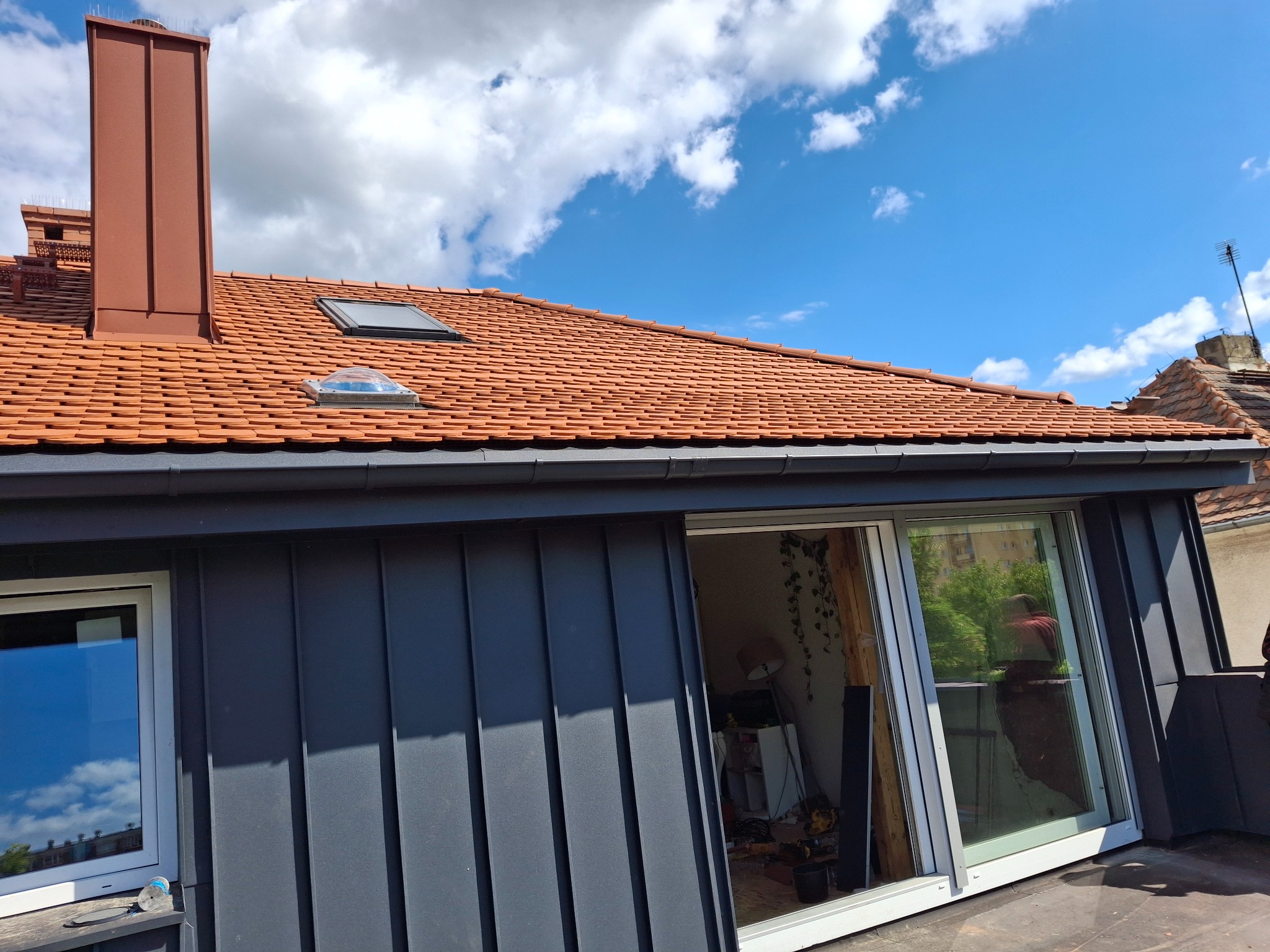 Czerwona dachówka ceramiczna z oknami dachowymi i kominem na tle błękitnego nieba z chmurami. Widoczne obróbki blacharskie i fragment elewacji budynku.