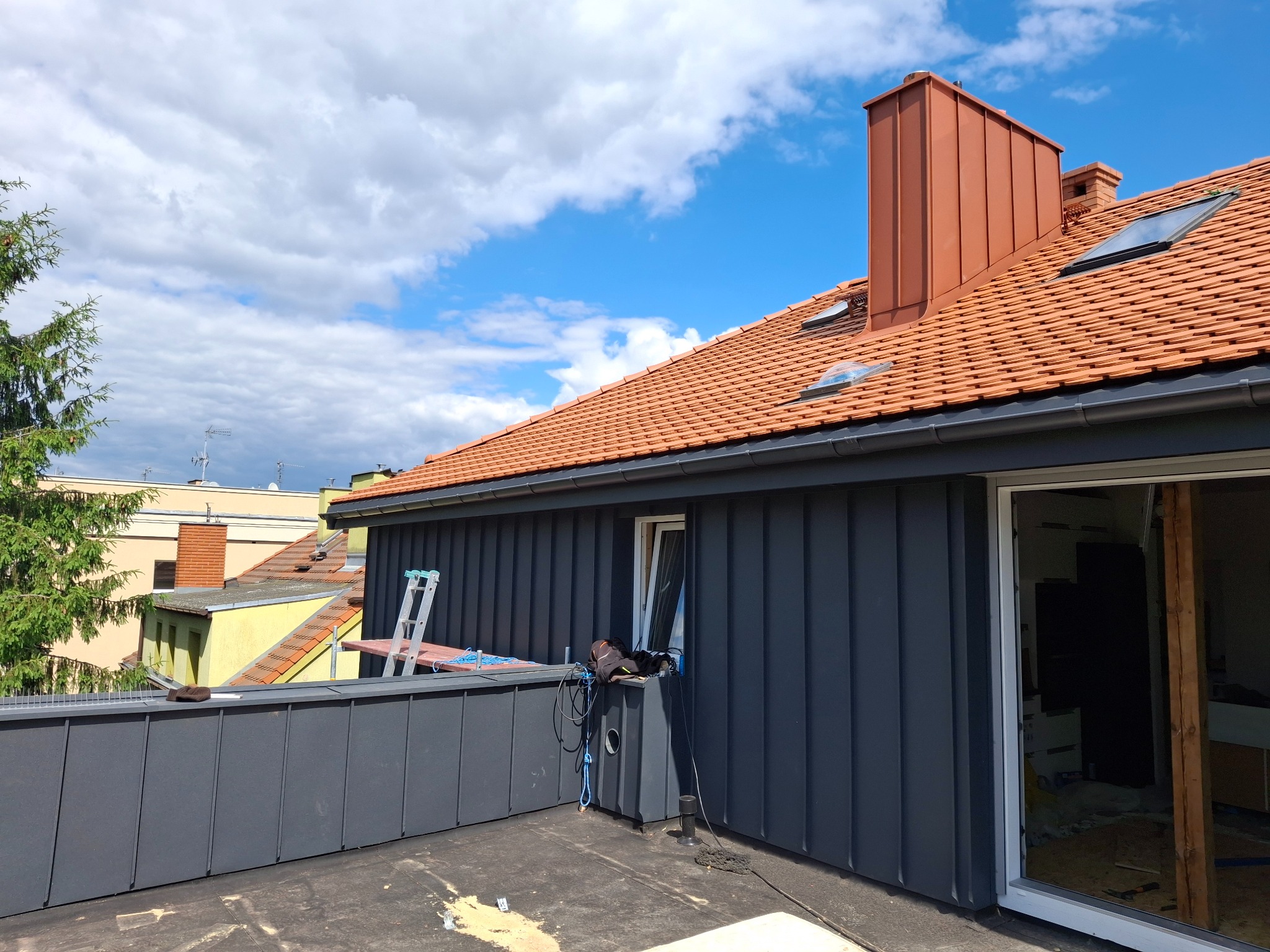Czerwona dachówka z lukarnami i kominem z blachy na tle błękitnego nieba. Widoczna szara obróbka blacharska i drabina oparta o ścianę budynku.