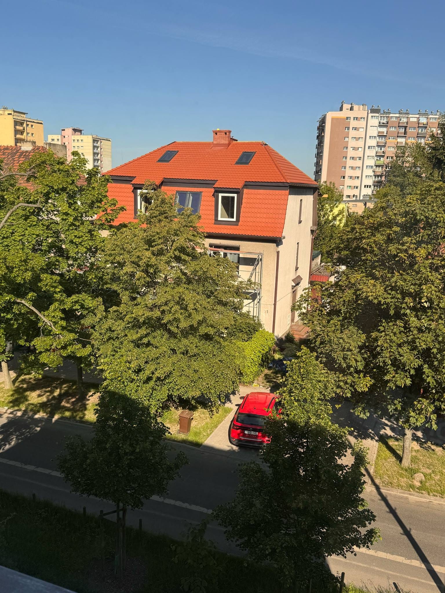 Dom z czerwoną dachówką i oknami dachowymi, otoczony zielenią drzew, z czerwonym samochodem na podjeździe. W tle bloki mieszkalne i błękitne niebo.