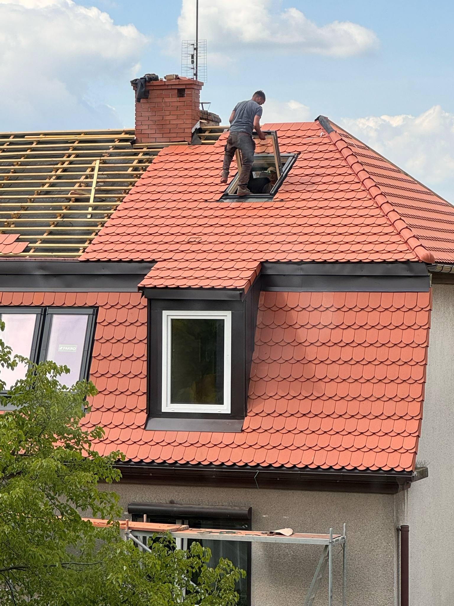 Czerwony dach z dachówką karpiówką w trakcie montażu okna dachowego. Widoczna więźba dachowa i komin z cegły. Praca na wysokości.