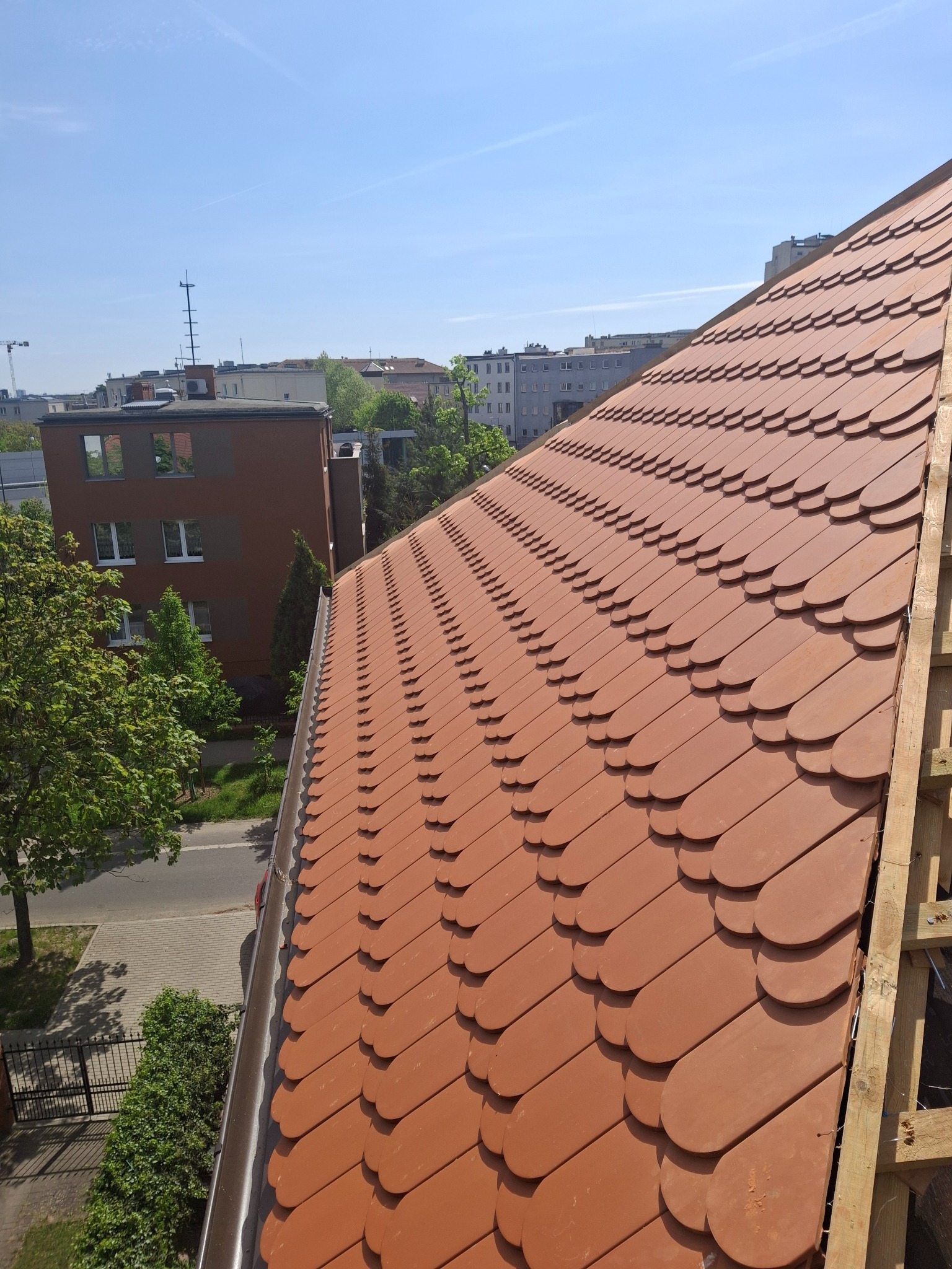 Dachówka karpiówka w odcieniach terakoty, ułożona na dachu budynku miejskiego. Widoczny fragment rynny i drewniana konstrukcja. W tle zabudowa miejska i błękitne niebo.