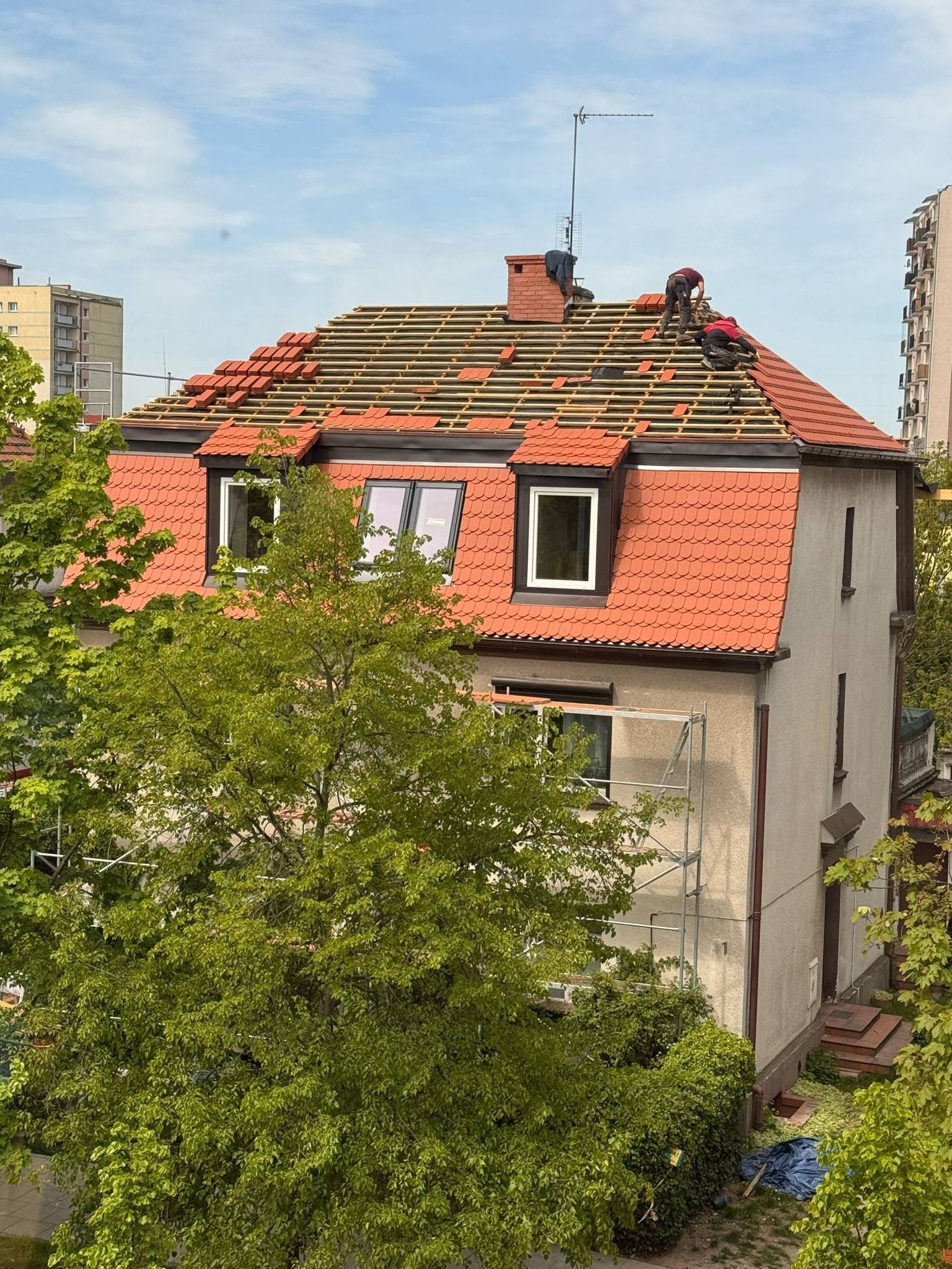Dwóch dekarzy pracuje na dachu domu z czerwonej dachówki, układając nowe pokrycie. Widoczne rusztowania i zieleń drzew. Ujęcie z góry, w tle budynki mieszkalne.