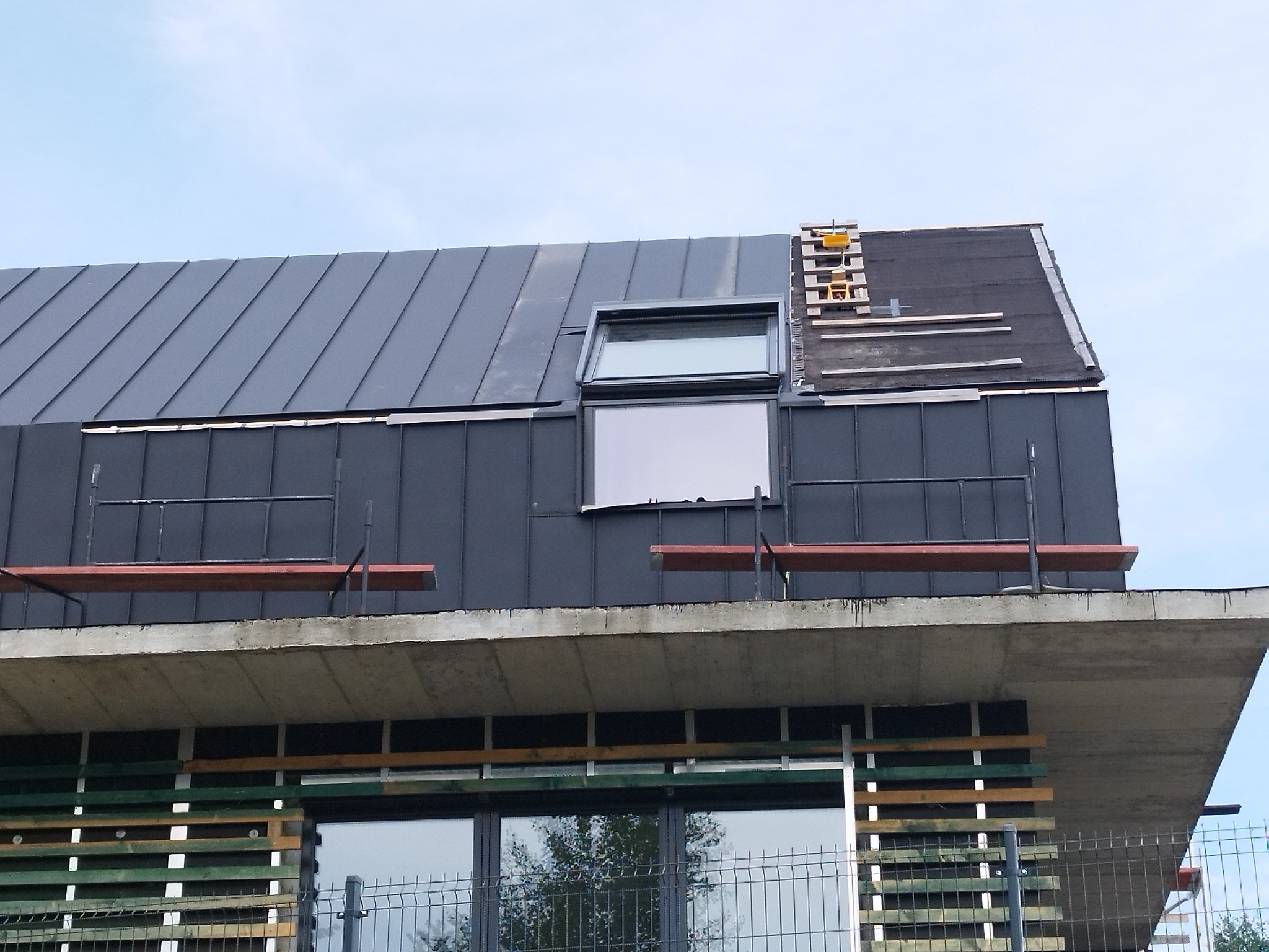 Nowoczesny dom w trakcie budowy z szarą blachą na dachu i oknem dachowym. Widoczne rusztowania i częściowo odkryta konstrukcja dachu.