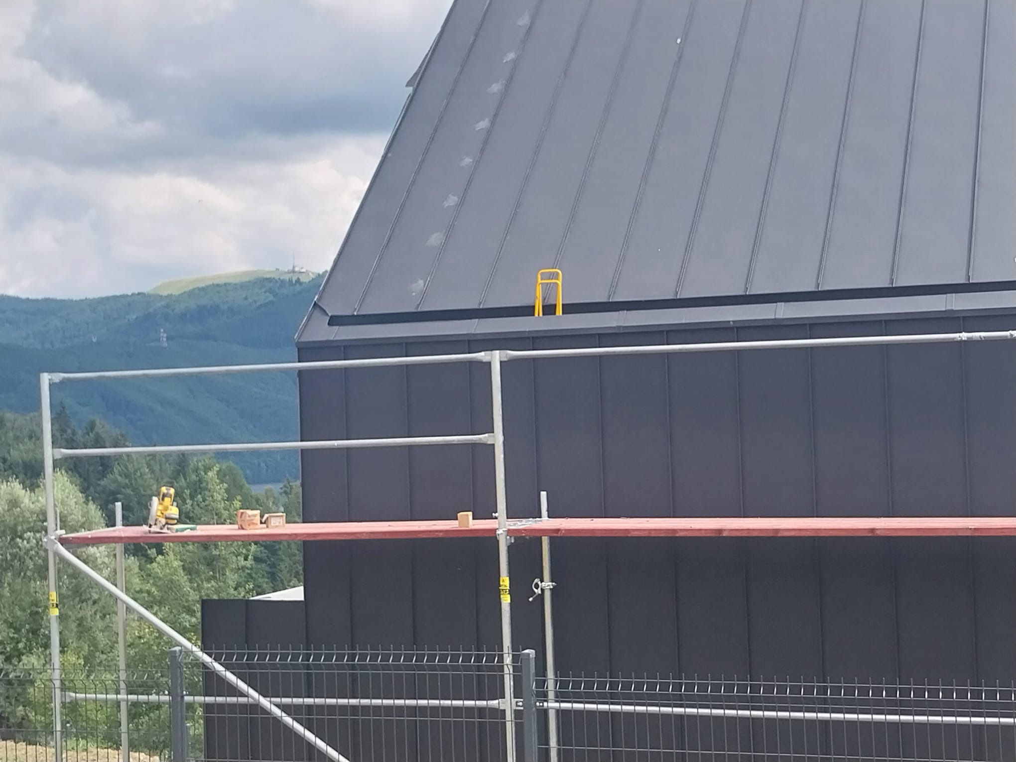 Szare poszycie dachu z blachy na tle górskim, widoczne rusztowanie z narzędziami, żółty element na dachu. Ujęcie z dołu, częściowy widok ogrodzenia.