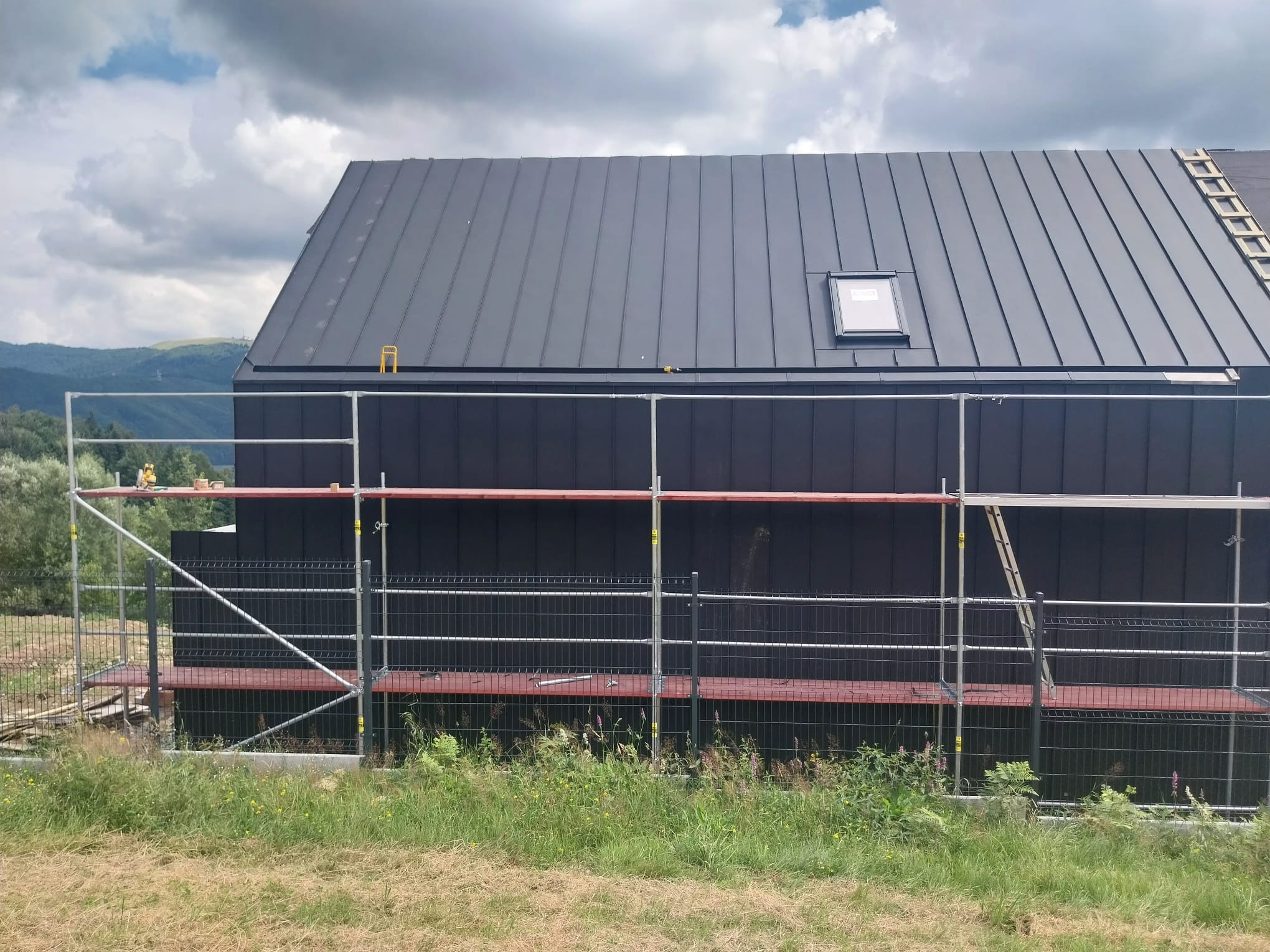 Czarny, blaszany dach jednospadowy z oknem dachowym, widoczny ze rusztowania na tle gór i pochmurnego nieba. Prace dekarskie w toku.