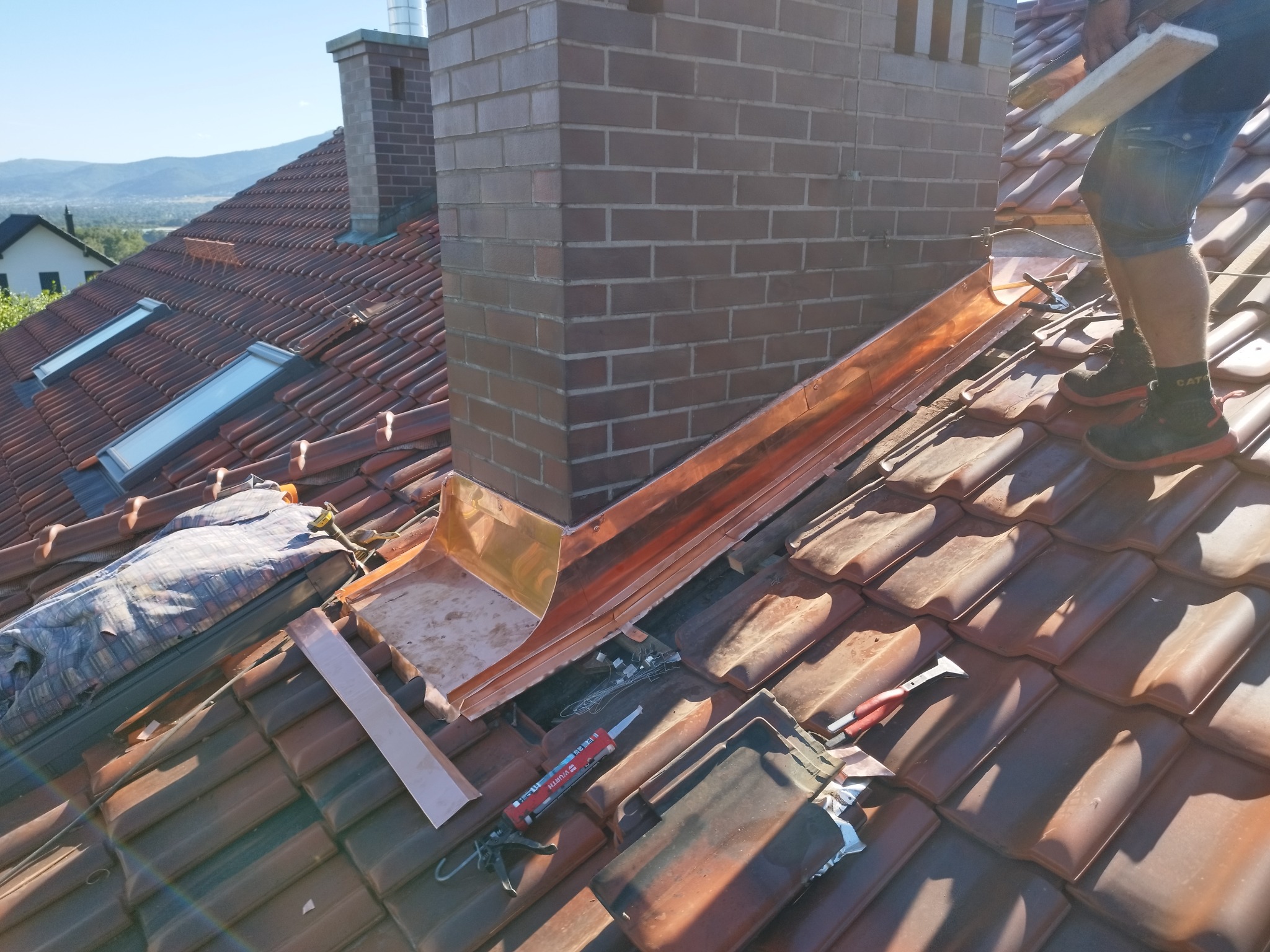 Montaż obróbki blacharskiej komina z miedzianej blachy na dachu z dachówki ceramicznej. Widoczne narzędzia i fragment sylwetki dekarza. Słoneczny dzień.