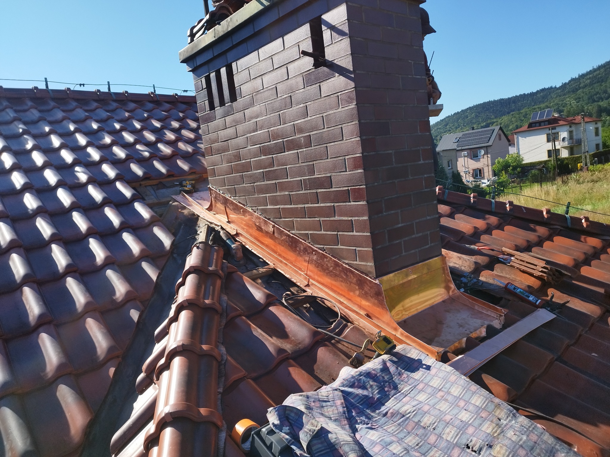 Ceglany komin z miedzianą obróbką blacharską na tle brązowej dachówki i błękitnego nieba. Widoczne narzędzia i fragment płachty zabezpieczającej.