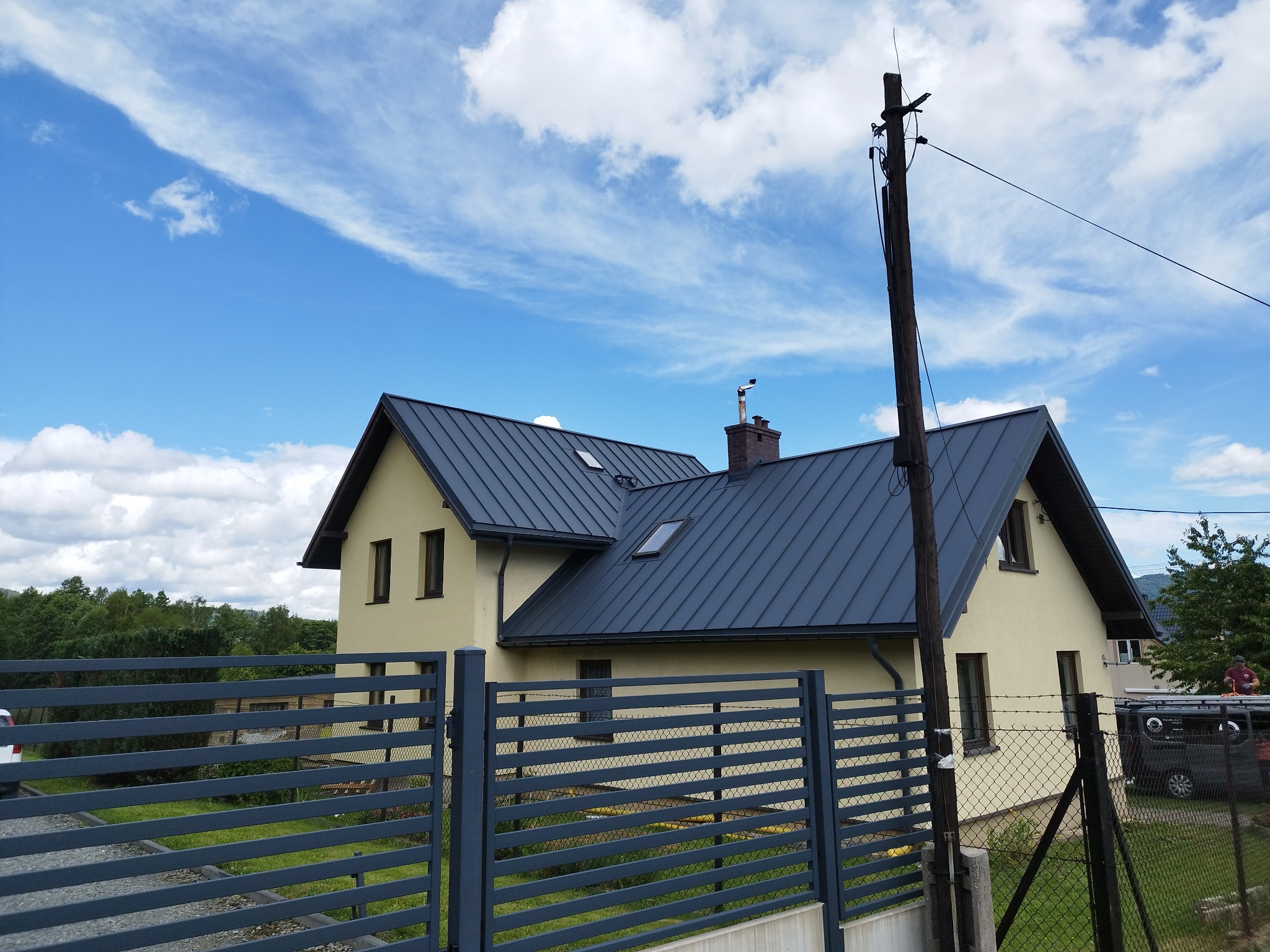 Dom jednorodzinny z ciemnoszarym dachem z blachy na tle błękitnego nieba z chmurami. Widoczny fragment ogrodzenia i słup elektryczny.