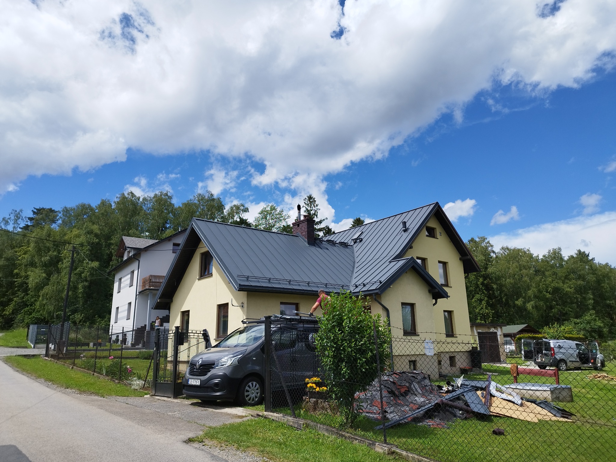 Żółty dom jednorodzinny z nowym, grafitowym dachem z blachy na tle błękitnego nieba z chmurami. Na pierwszym planie samochód dostawczy i pozostałości po pracach dekarskich.