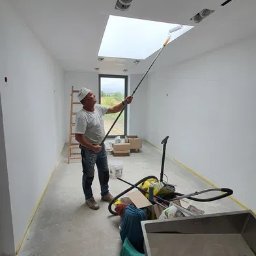 Zakład Remontowo-Budowlany Kolorek - Malarz w białej czapce maluje sufit wałkiem na długim kiju w jasnym, długim pomieszczeniu z oknem i drabinką. Widoczne narzędzia i akcesoria malarskie.