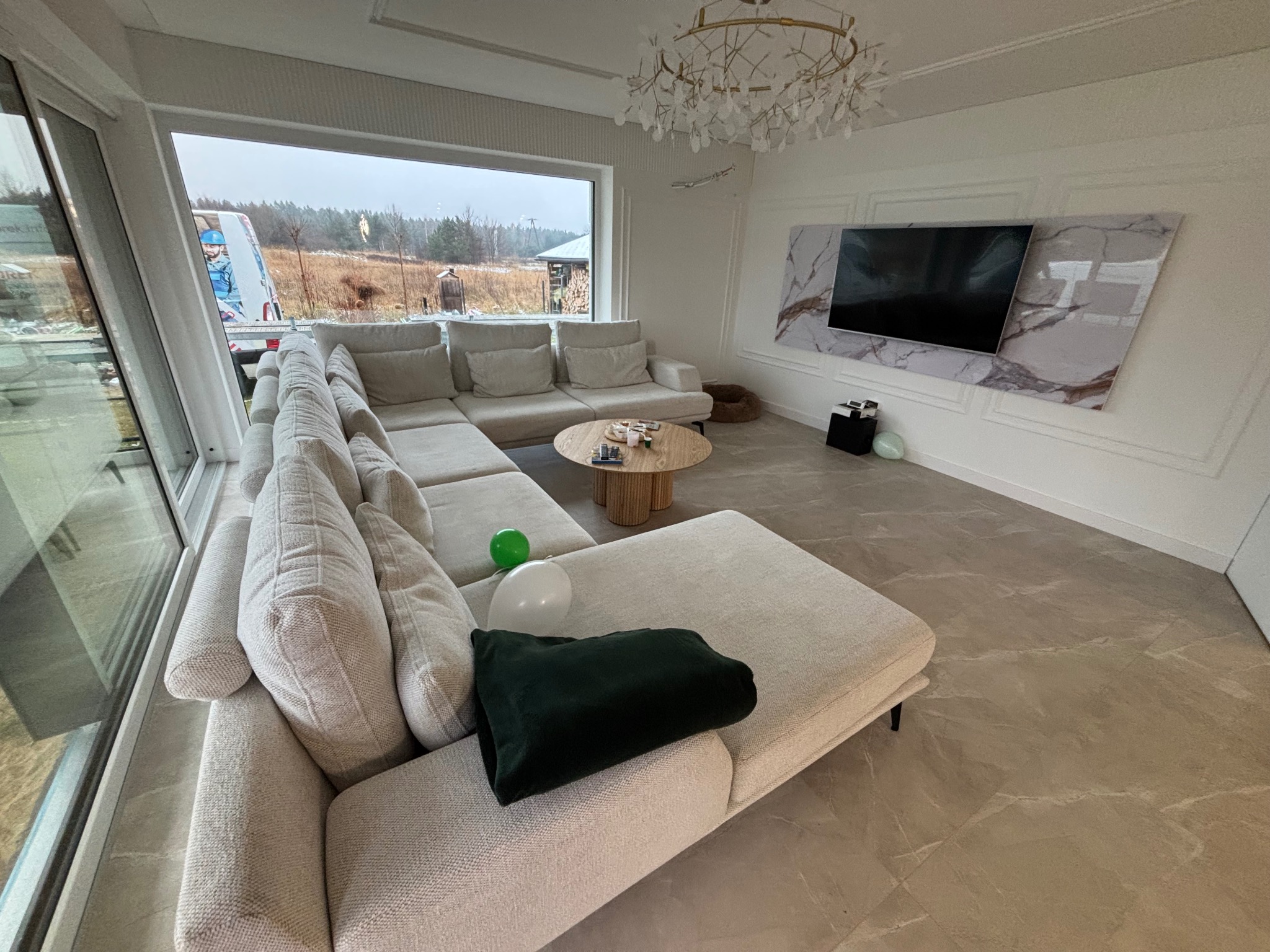 Jasny salon z narożnikiem, podwieszanym sufitem i panelem TV z marmurowym wzorem. Widok na zewnątrz przez duże okno. Nowoczesne wykończenie wnętrza.