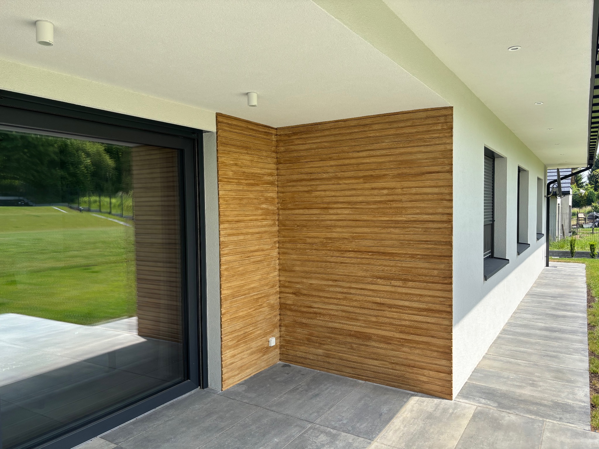 Wykończona elewacja domu z drewnianą okładziną, widok z zewnątrz. Szara kostka brukowa na tarasie, okna z czarnymi ramami, biała ściana. Nowoczesny design.