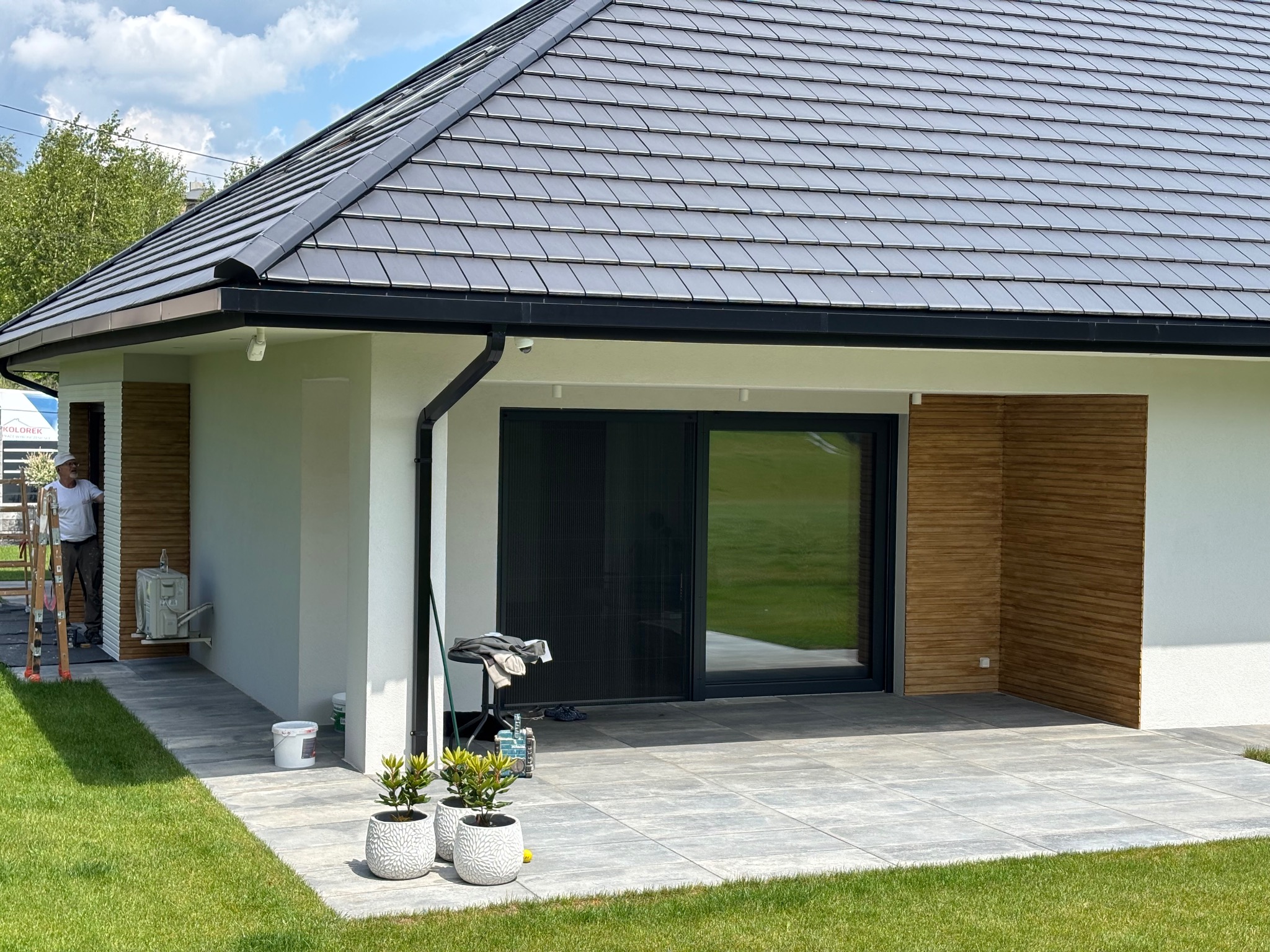 Nowoczesny dom jednorodzinny po malowaniu elewacji, z szarą dachówką i elementami drewnianymi. Widoczne narzędzia malarskie i pracownik w tle.