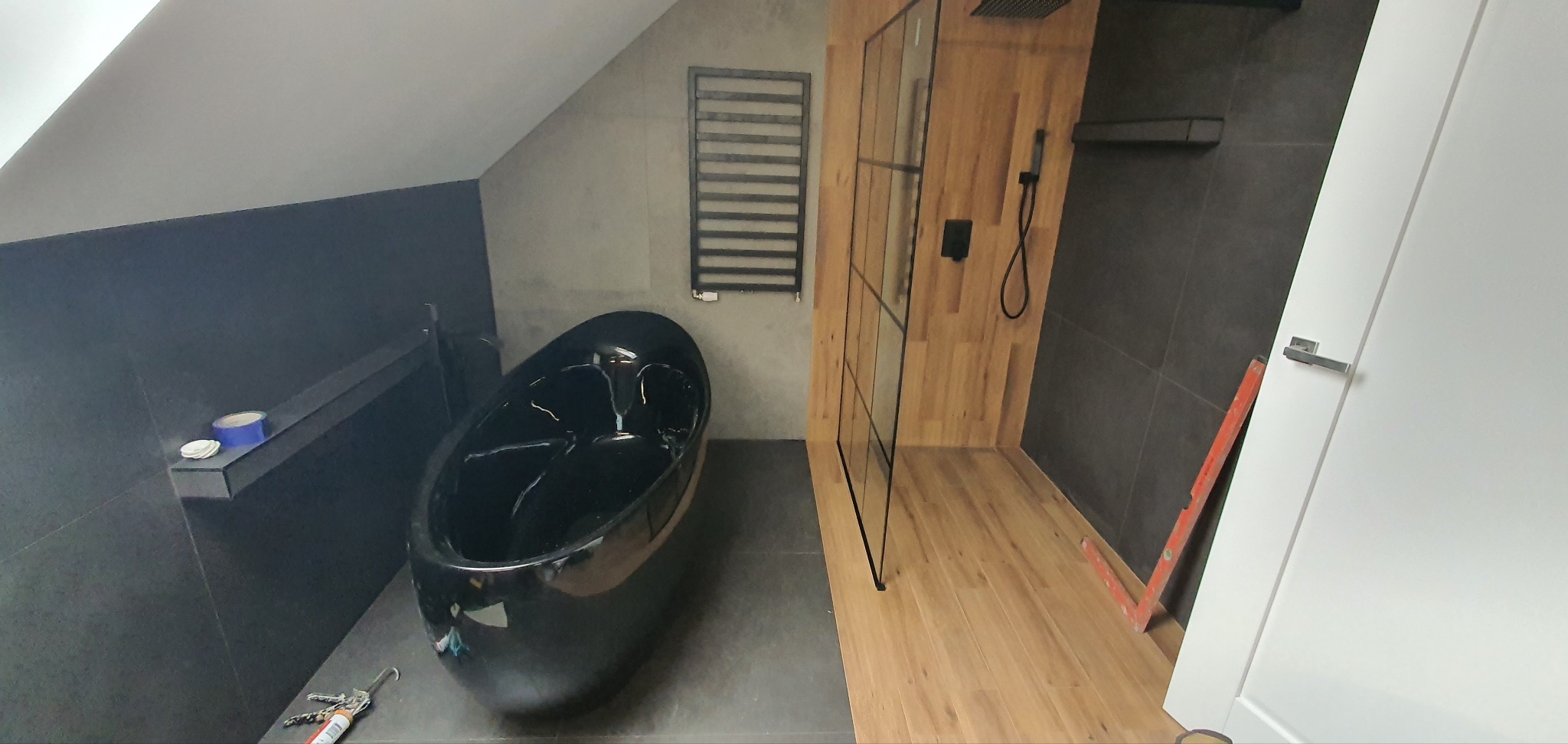 Nowoczesna łazienka na poddaszu z czarną wanną wolnostojącą, drewnianą podłogą i szklaną kabiną prysznicową. Widoczne narzędzia i poziomica.