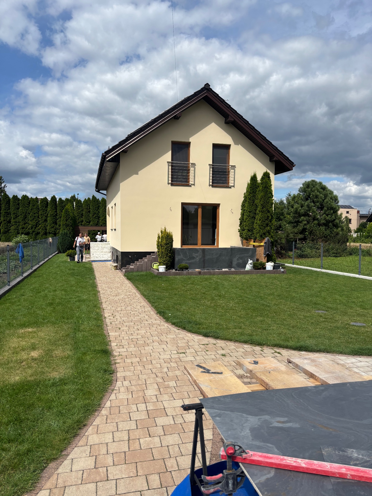 Jasna elewacja domu jednorodzinnego z balkonami, widok z ogrodu z brukowanym chodnikiem. W tle grupa osób i wysokie tuje. W kadrze narzędzia i płyty.