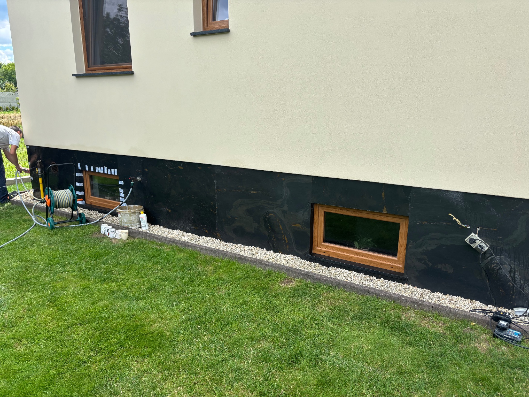 Przygotowanie elewacji budynku do malowania: zabezpieczona dolna część ściany, widoczne okna i narzędzia pracy na trawniku.