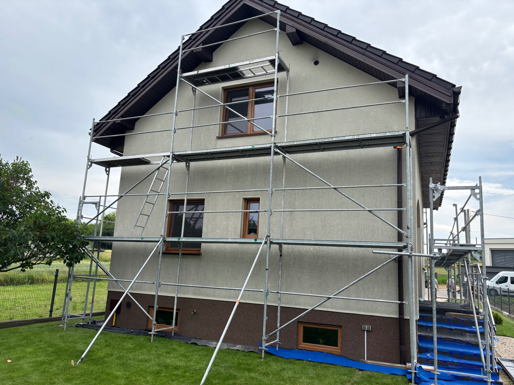 Dom jednorodzinny w trakcie prac elewacyjnych, otoczony rusztowaniem. Widoczne okna z brązowymi ramami i fragment ogrodu z zielonym trawnikiem.