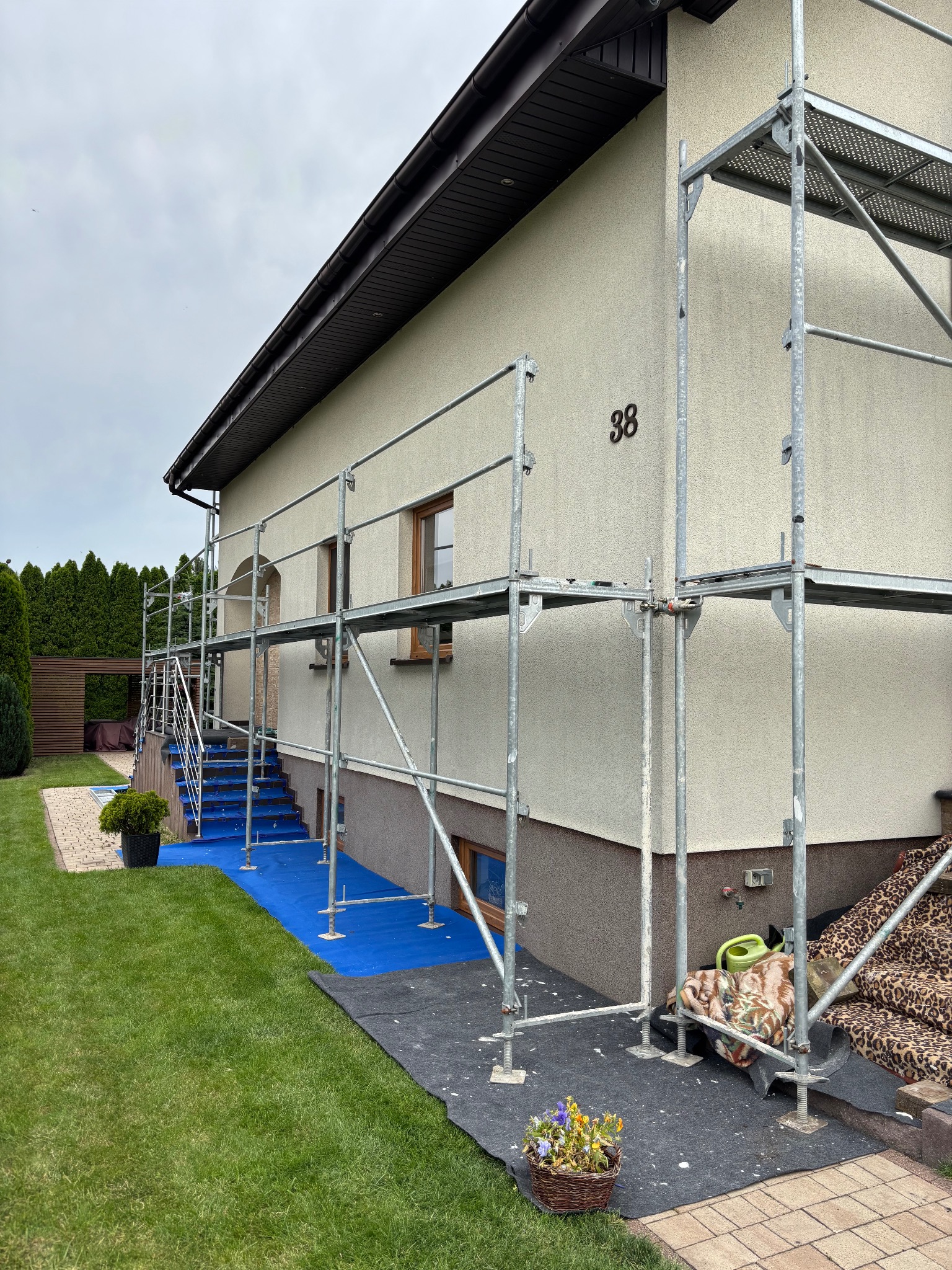 Dom jednorodzinny w trakcie prac elewacyjnych. Widoczne rusztowania, zabezpieczenia na schodach i trawniku. Numer domu '38' na jasnej fasadzie.