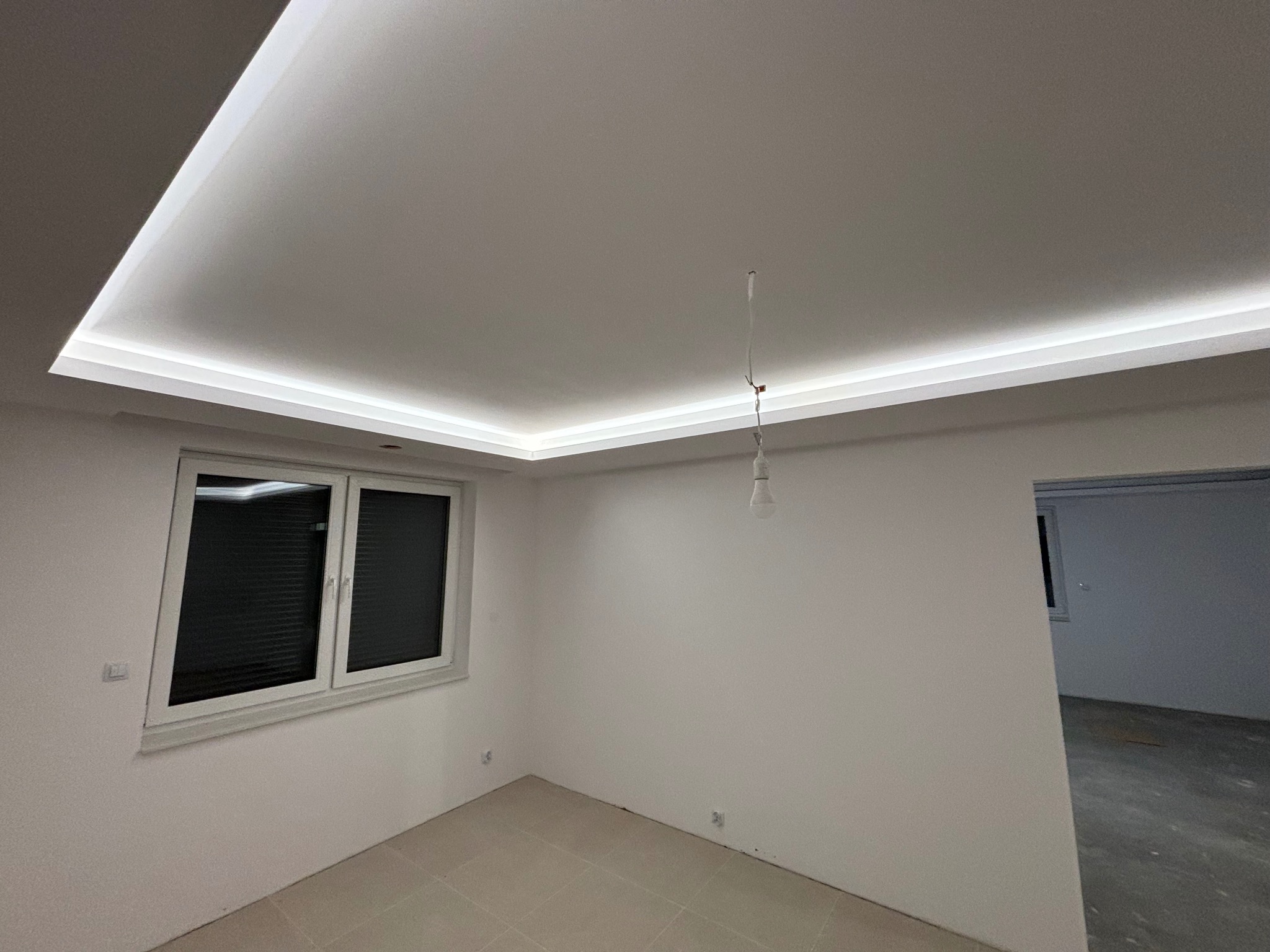 Białe wnętrze z podwieszanym sufitem i oświetleniem LED. Widoczne okno z żaluzjami i fragment sąsiedniego pomieszczenia. Minimalistyczny design.