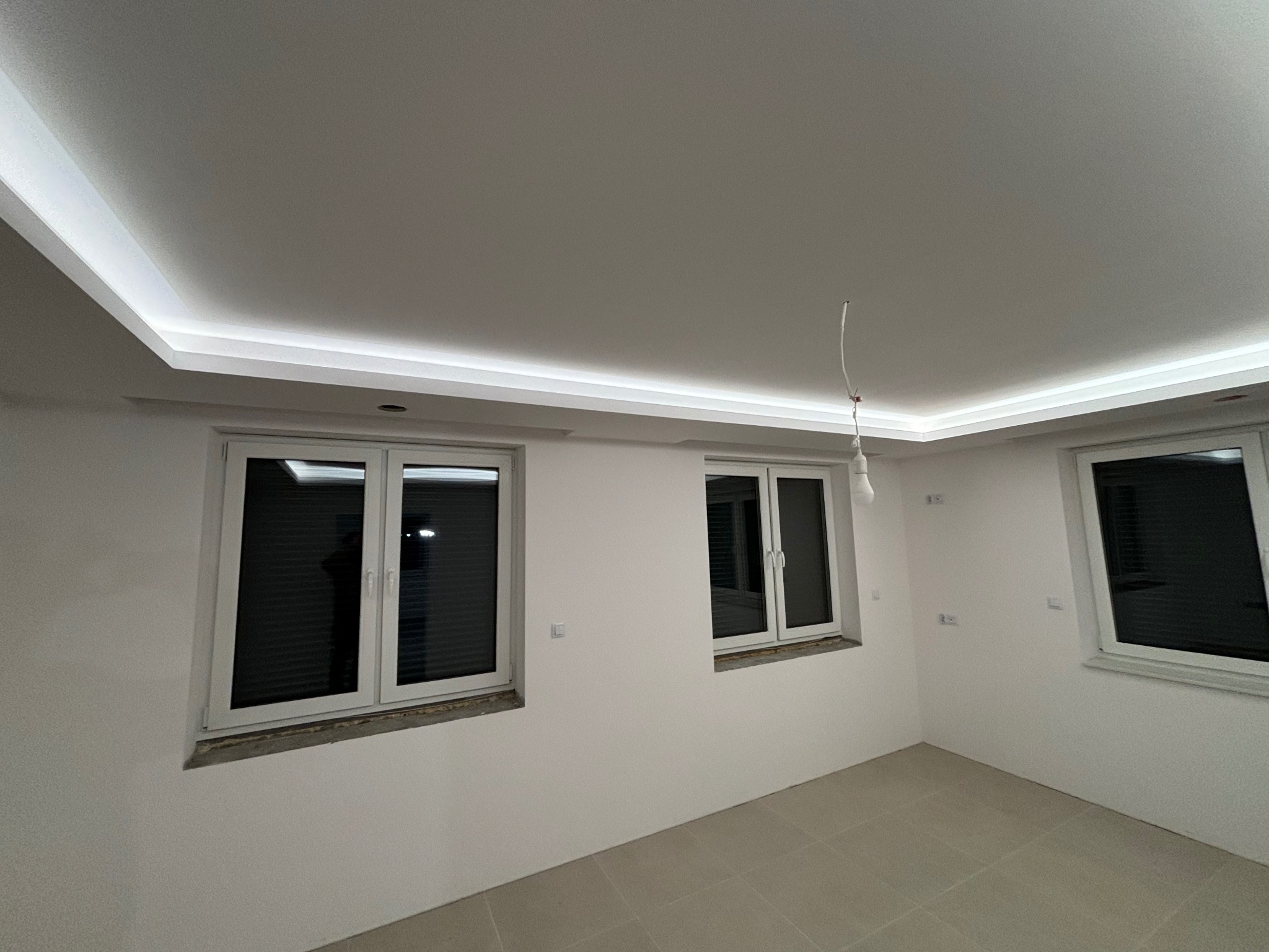 Białe wnętrze po remoncie z trzema oknami i podświetlanym sufitem. Widoczne gładkie ściany i płytki na podłodze. W tle wiszący kabel od lampy.