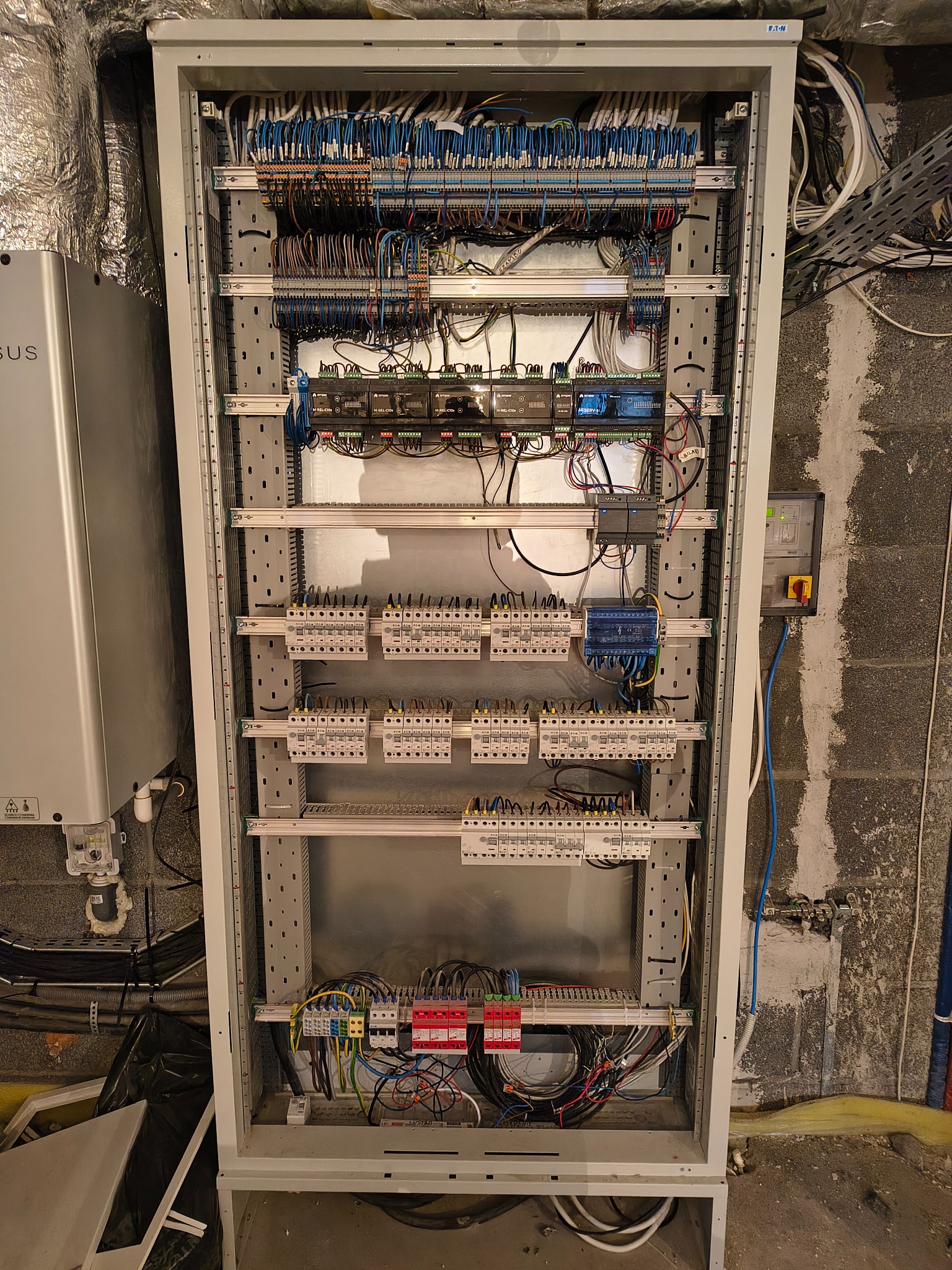 Rozdzielnia elektryczna w metalowej obudowie z licznymi przewodami, bezpiecznikami i modułami sterującymi. Widoczne kable w różnych kolorach i elementy instalacji.