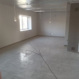 KAP-BUD - Jasne, wykafelkowane wnętrze w trakcie wykańczania. Widoczne kable, surowy sufit i okno. Minimalistyczny styl, neutralna kolorystyka. Etap prac wykończeniowych.