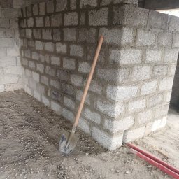 KAP-BUD - Ściana z betonu komórkowego w trakcie budowy, widoczna zaprawa i narzędzie - łopata oparta o ścianę. W tle widoczne elementy konstrukcyjne budynku.