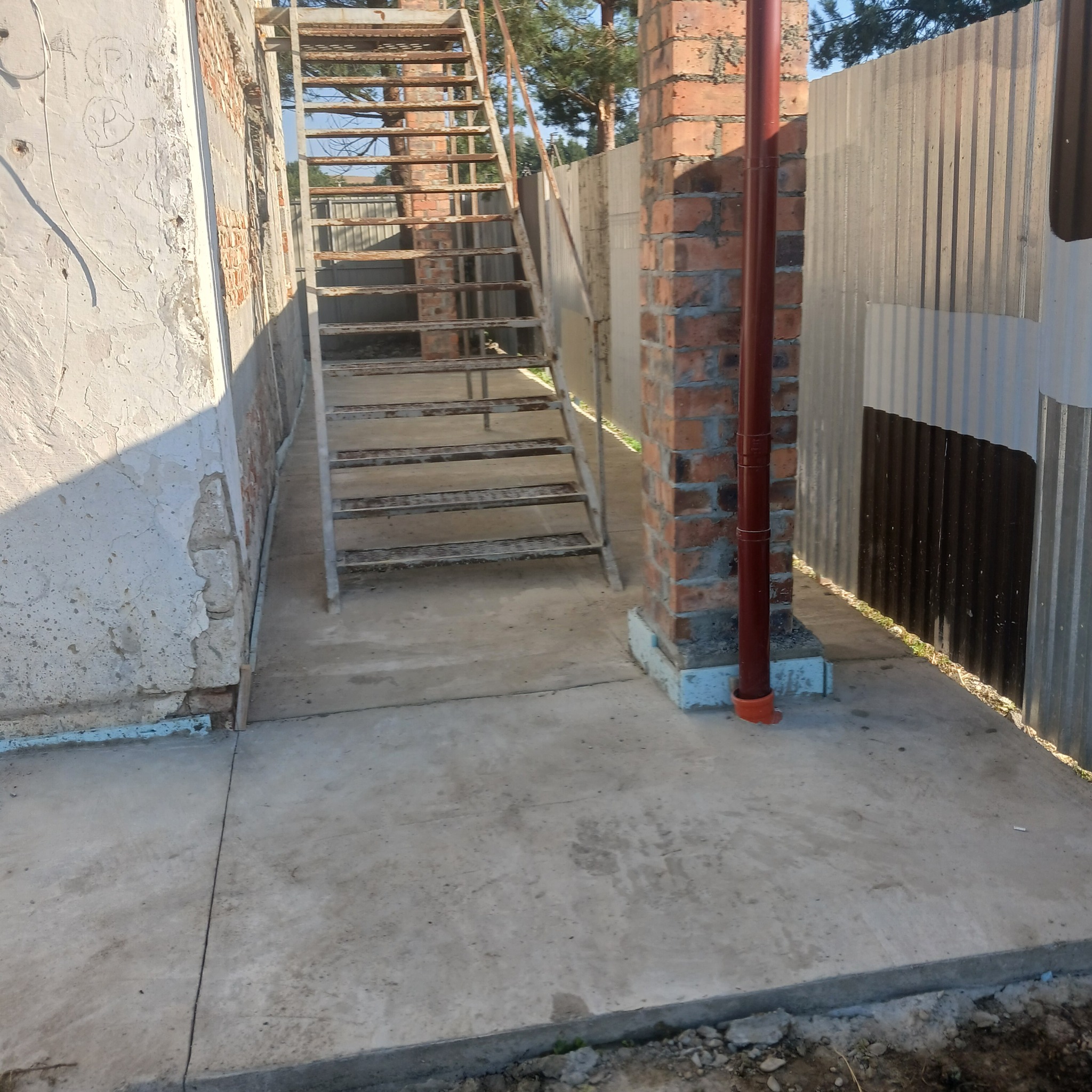 Betonowa wylewka pod schodami w Przemyślu, widok z góry. Metalowe schody, ceglany filar i blaszane ogrodzenie w tle. Świeża, jasnoszara powierzchnia betonu.