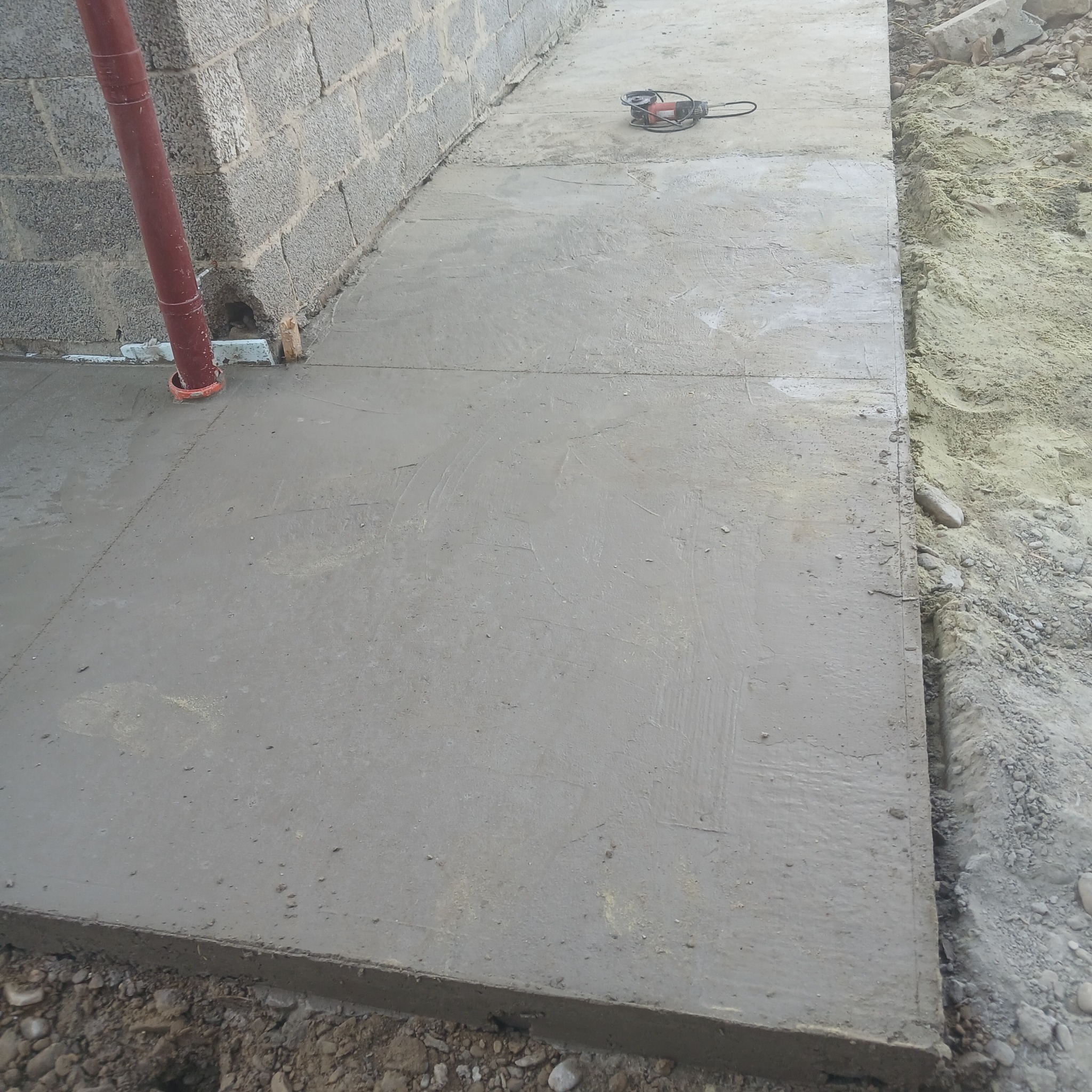 Świeżo wylana, szara betonowa wylewka chodnika przy budynku z szarego bloczka. Widoczna rura spustowa i szlifierka kątowa. Krawędź chodnika styka się z ziemią.