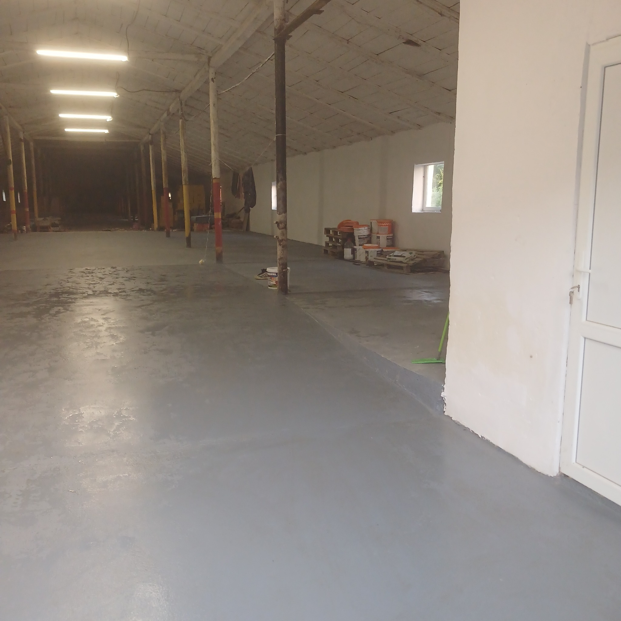 Szara, gładka posadzka w hali magazynowej po wylaniu wylewki samopoziomującej. Widoczne puszki z farbą i narzędzia w tle. Oświetlenie jarzeniowe.