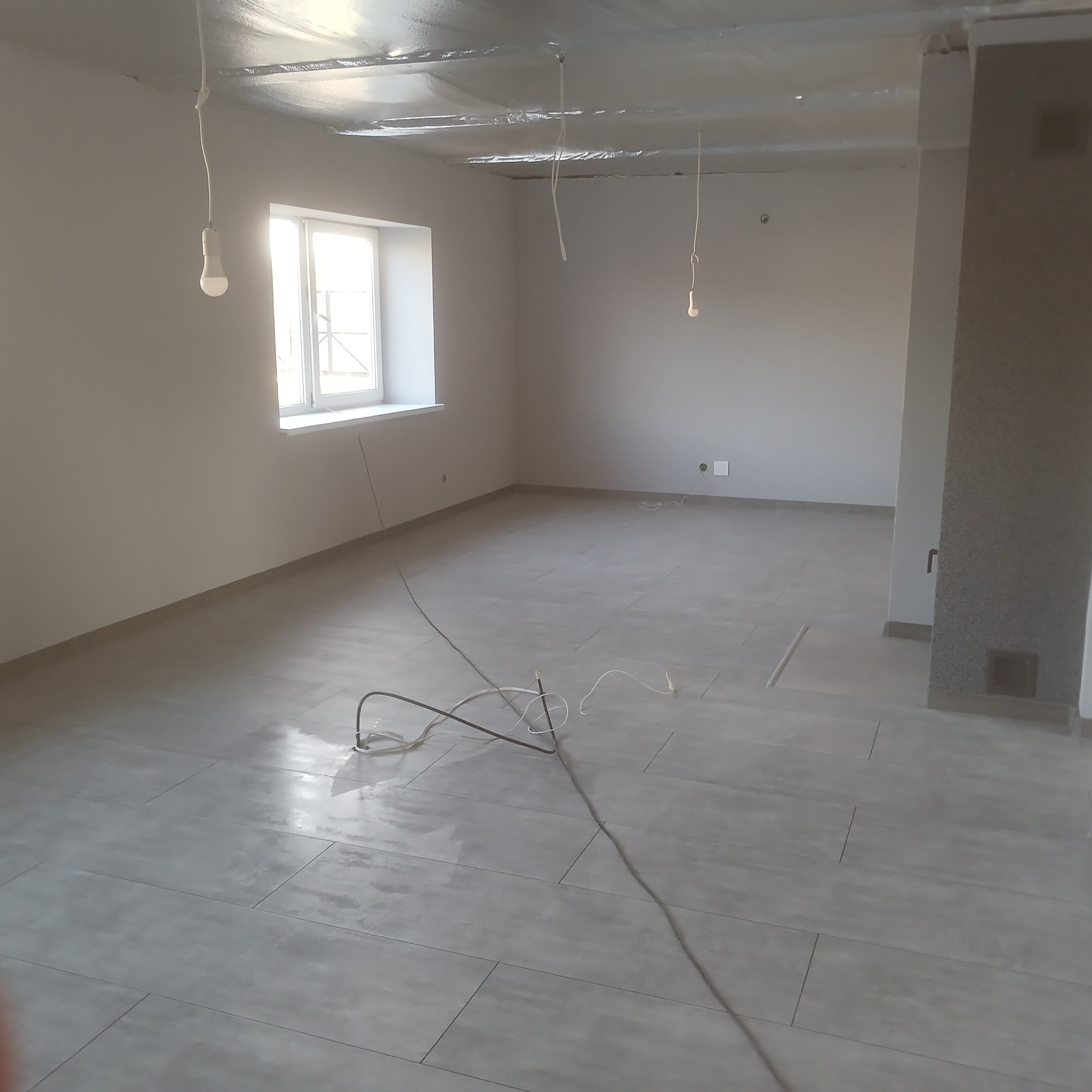 Jasne, wykafelkowane wnętrze w trakcie wykańczania. Widoczne kable, surowy sufit i okno. Minimalistyczny styl, neutralna kolorystyka. Etap prac wykończeniowych.