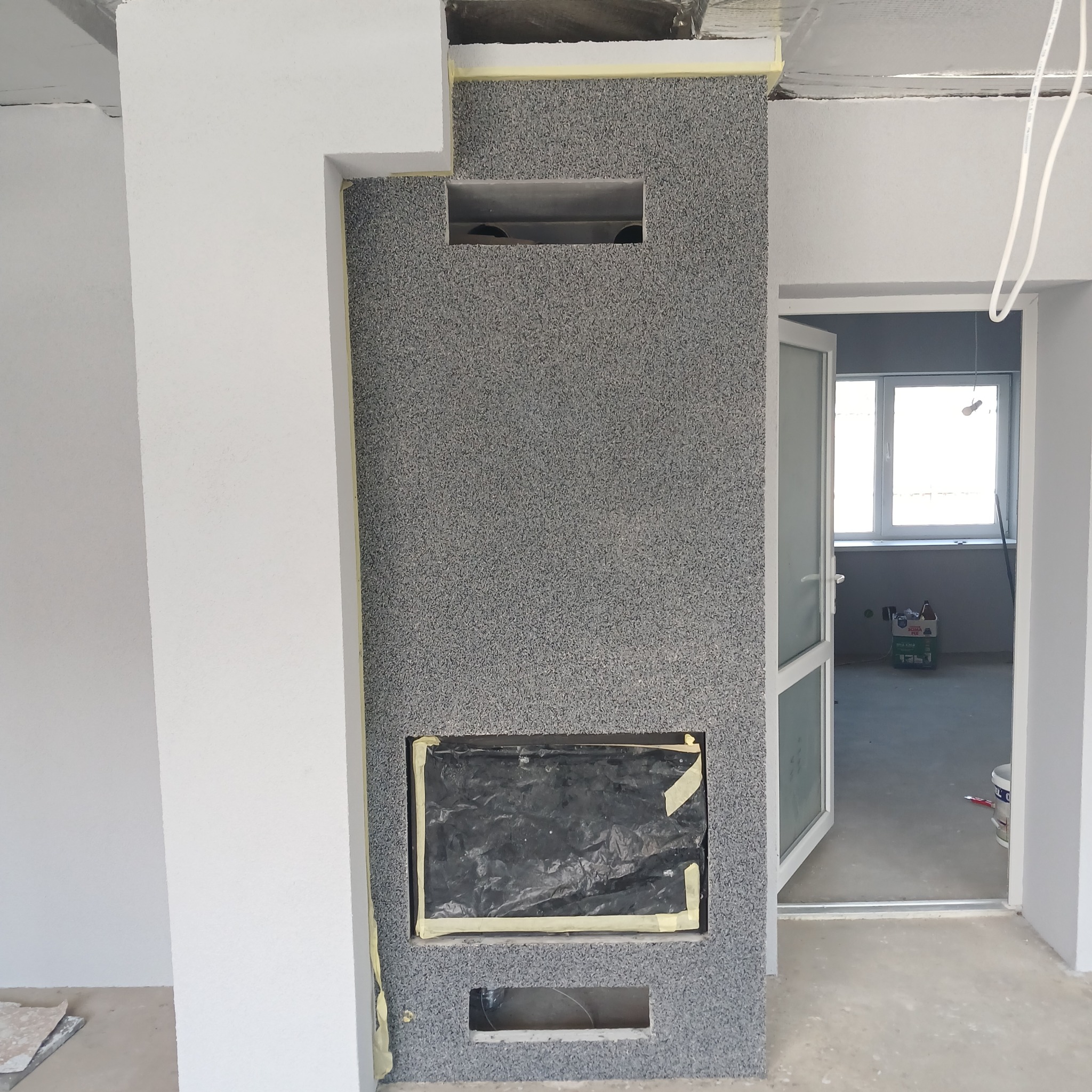Ściana z szarą, ziarnistą strukturą, z otworem na kominek zabezpieczonym folią i taśmą. Widoczne fragmenty białych ścian i otwarte drzwi do innego pomieszczenia.