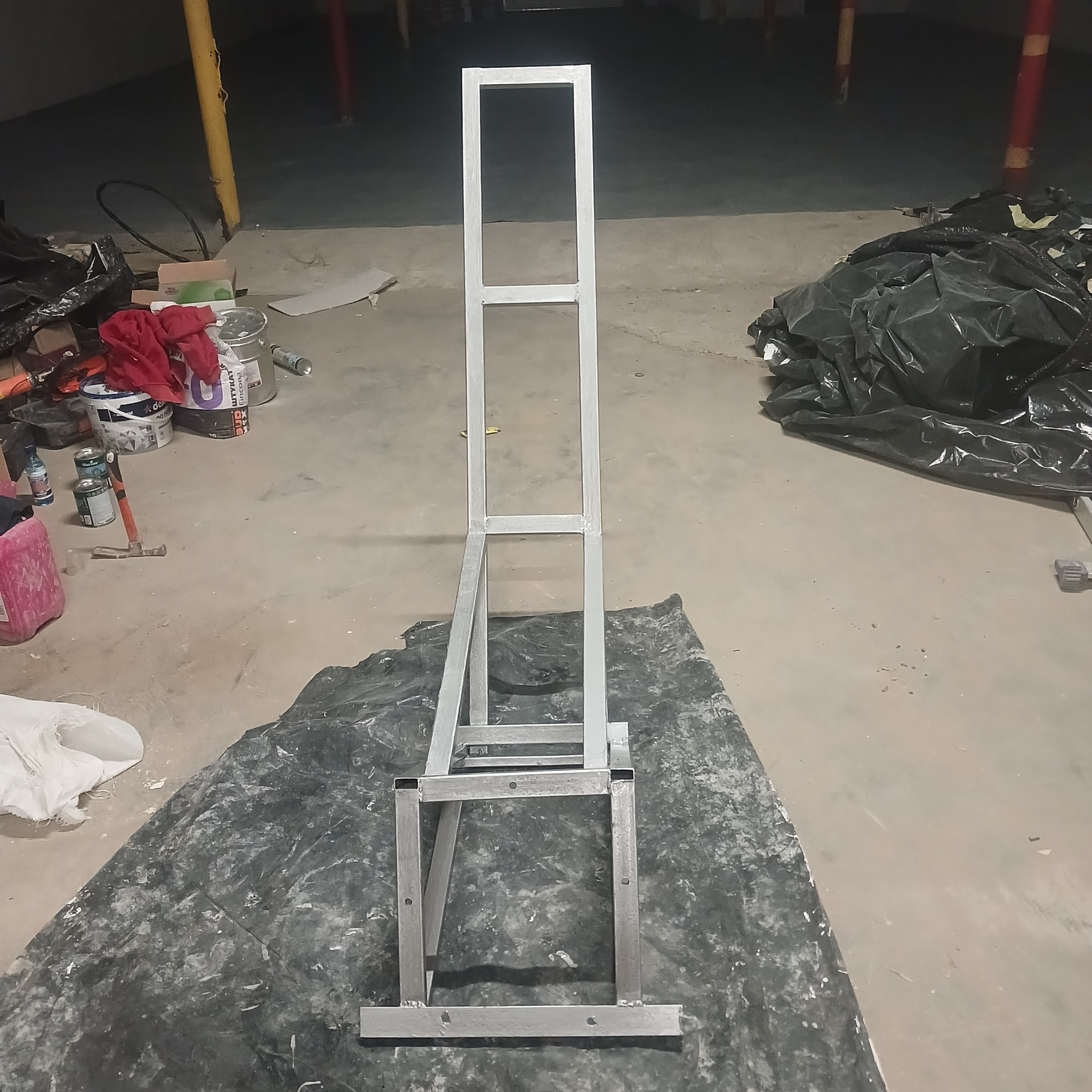 Metalowa konstrukcja w surowym wnętrzu, prawdopodobnie rama pod sufit napinany, widoczne narzędzia i farby w tle. Ujęcie z niskiej perspektywy.