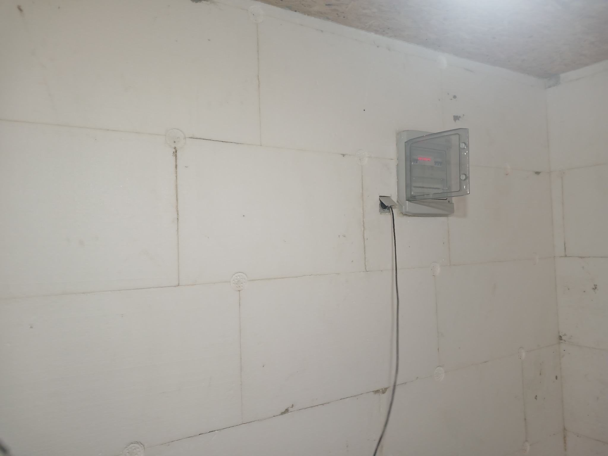 Ściana z białych płyt styropianowych z widoczną, otwartą skrzynką elektryczną i wystającym kablem. Widoczne łączenia płyt i elementy mocujące.