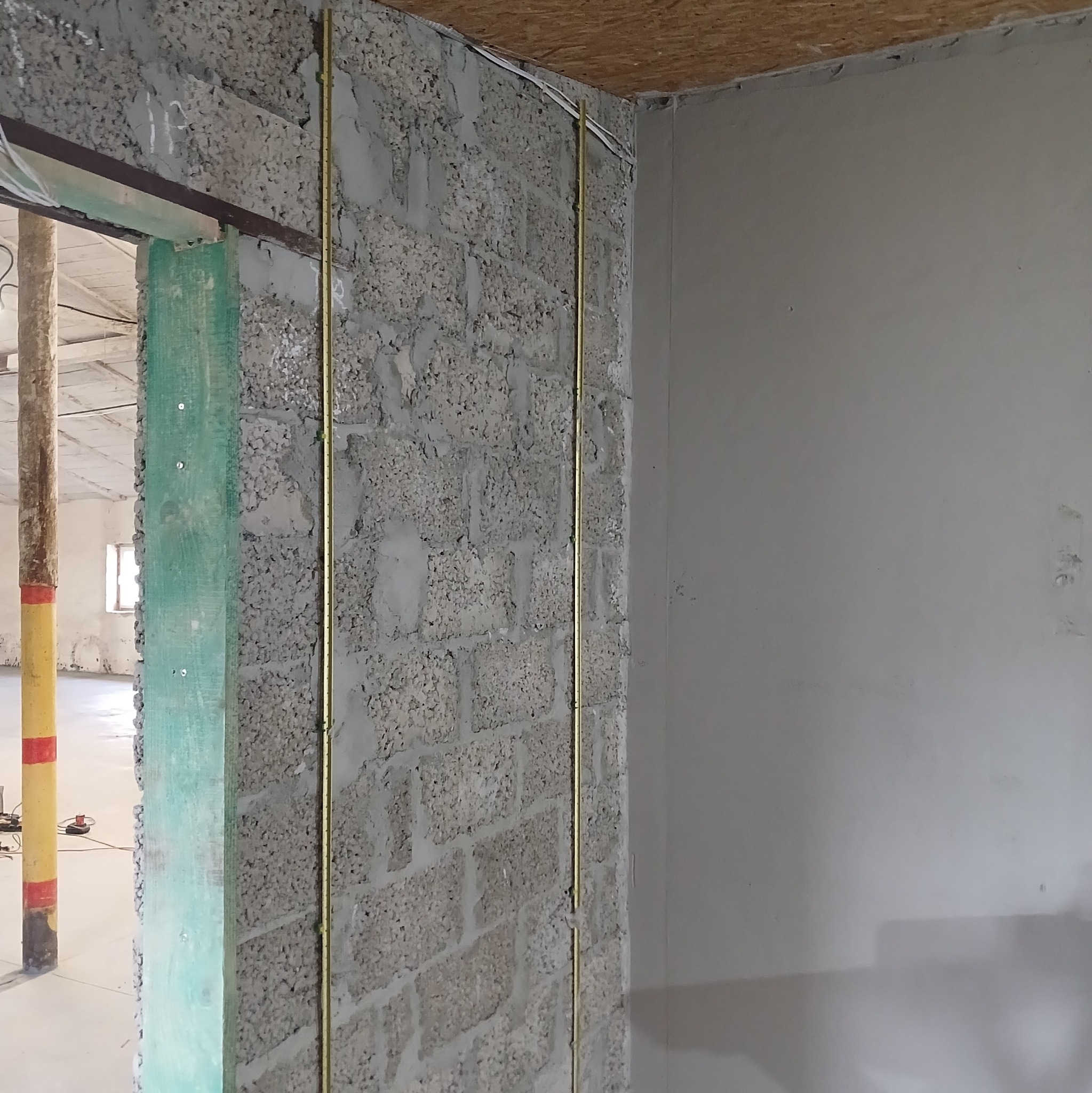 Ściana z betonu komórkowego z pionowymi rurkami instalacyjnymi, widok na surowy stan wykończenia wnętrza, fragment otworu drzwiowego z zieloną osłoną.