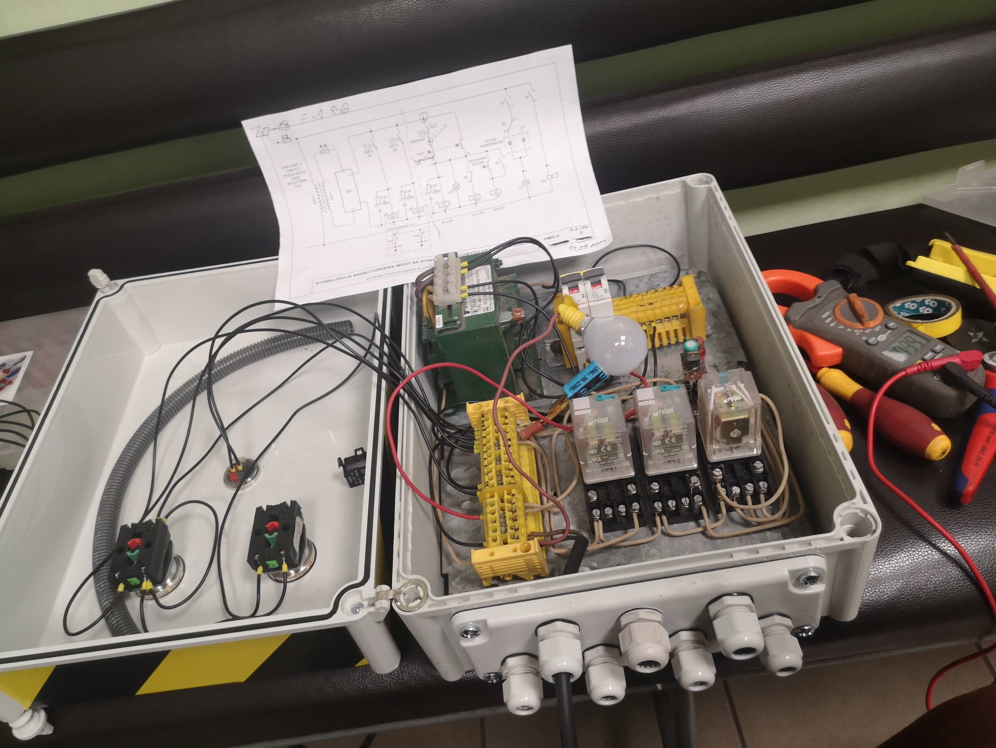 Otwarta skrzynka elektryczna z elementami instalacji, schematem i narzędziami. Widoczne przekaźniki, złączki, przewody i miernik. Praca w toku, detale instalacyjne.