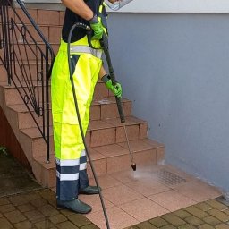 Burklin Marcin Burak - Pracownik w odzieży ochronnej czyści ciśnieniowo płytki chodnikowe przed schodami wejściowymi. Widoczny strumień wody i czyszczona powierzchnia.