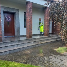 Burklin Marcin Burak - Pracownik w odblaskowym stroju czyści kostkę brukową przed domem za pomocą myjki ciśnieniowej. Widoczne schody wejściowe i fragment elewacji.