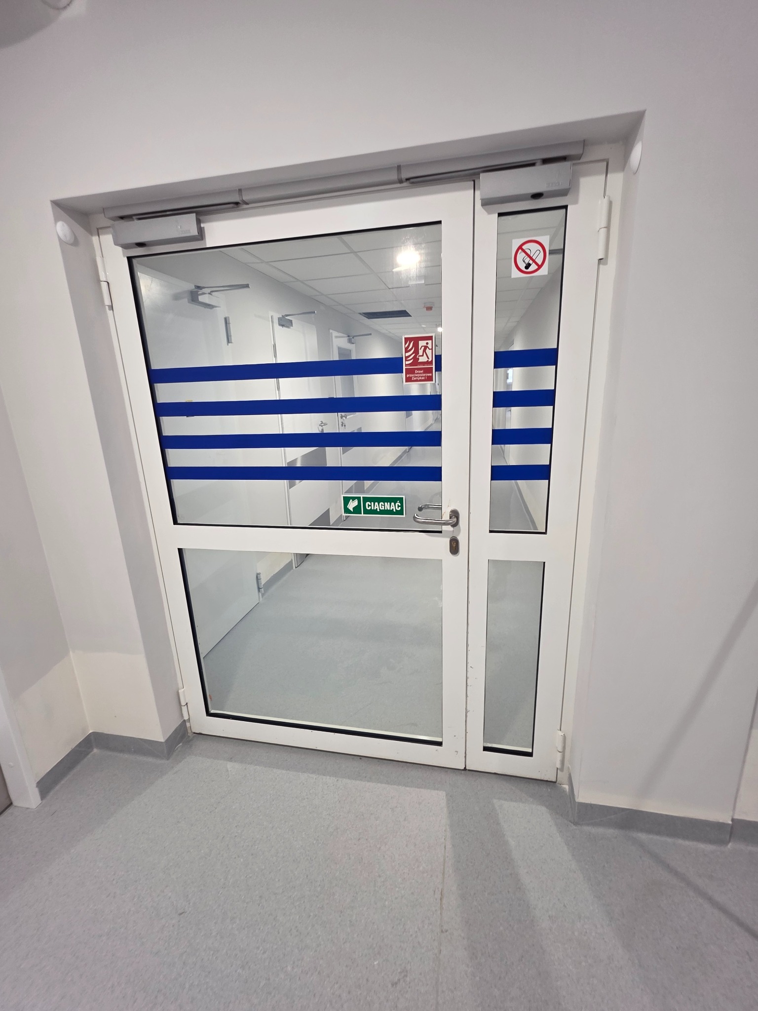 Białe, przeszklone drzwi z niebieskimi pasami i oznaczeniami bezpieczeństwa, prowadzące do czystego, sterylnego pomieszczenia z półkami. Widok z bliska, neutralne oświetlenie.