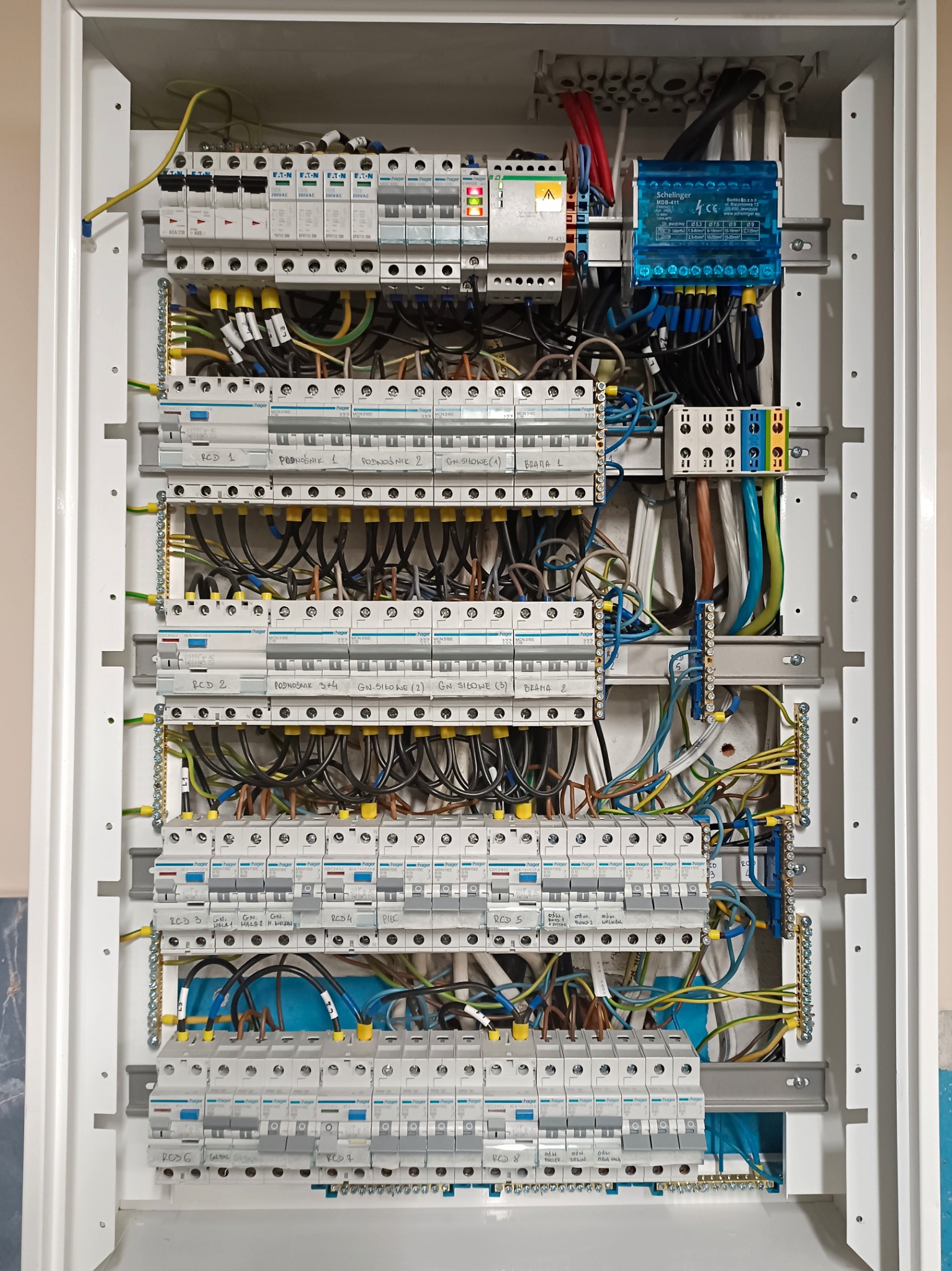 Rozdzielnica elektryczna w białej obudowie, z licznymi bezpiecznikami, przewodami i elementami instalacji, starannie ułożonymi i podłączonymi. Widoczne oznaczenia i etykiety.