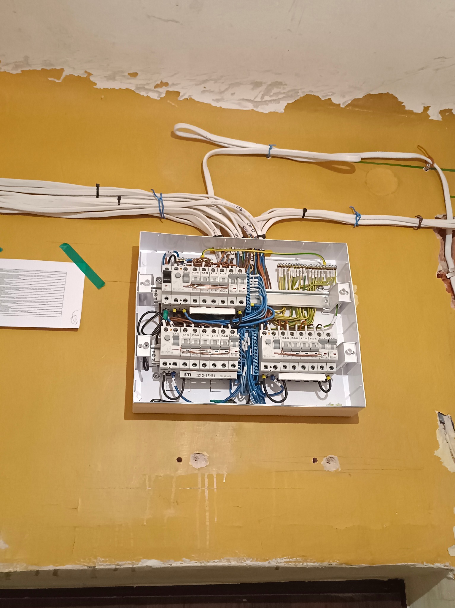 Rozdzielnica elektryczna z przewodami na żółtej ścianie w trakcie montażu w Ząbkach. Widoczne kable i elementy zabezpieczające. Ściana wymaga odświeżenia.