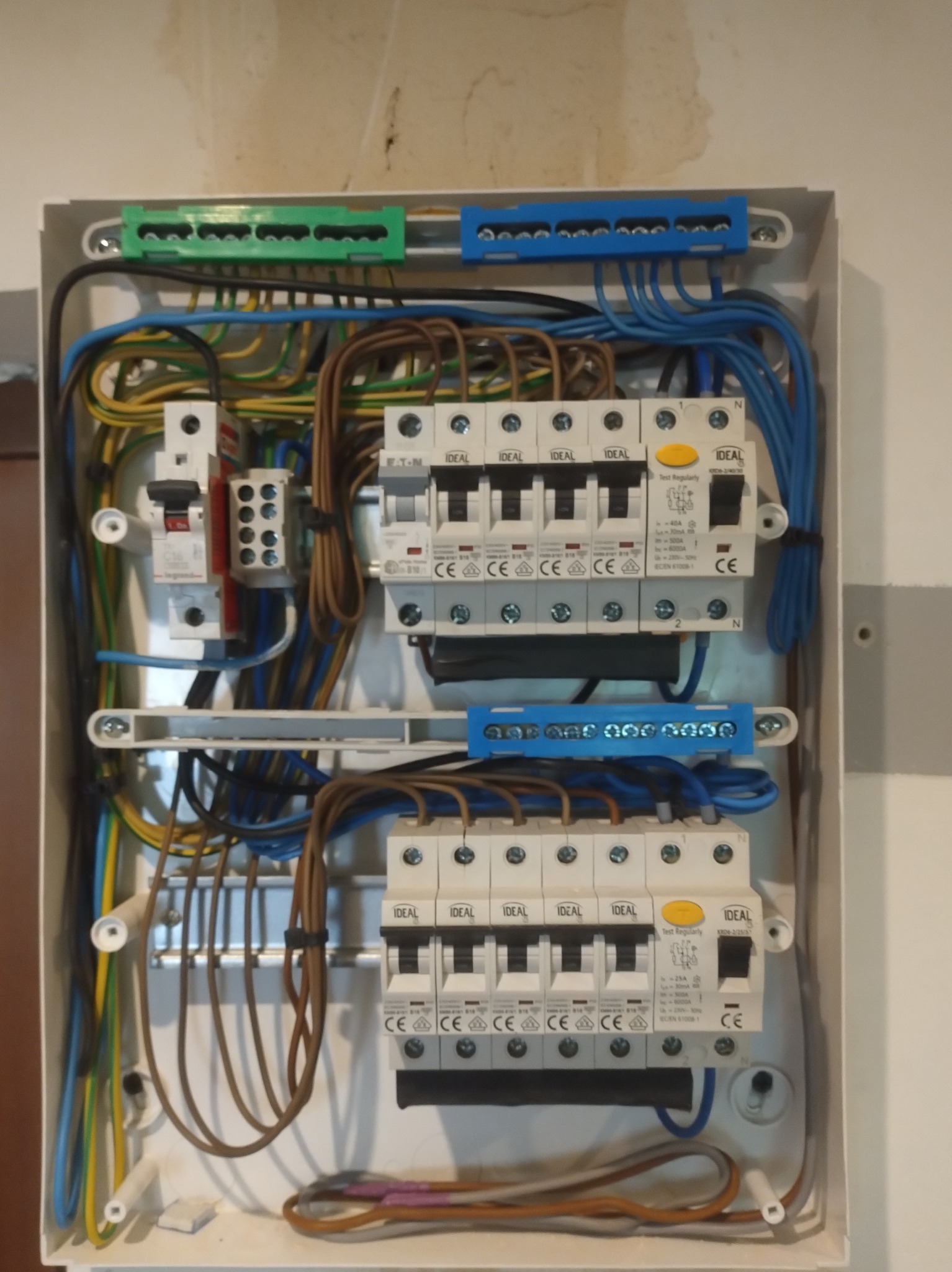 Rozdzielnica elektryczna z bezpiecznikami IDEAL, widoczne przewody w kolorach: niebieskim, brązowym, zielono-żółtym. Instalacja w białej obudowie, zamontowana na ścianie.