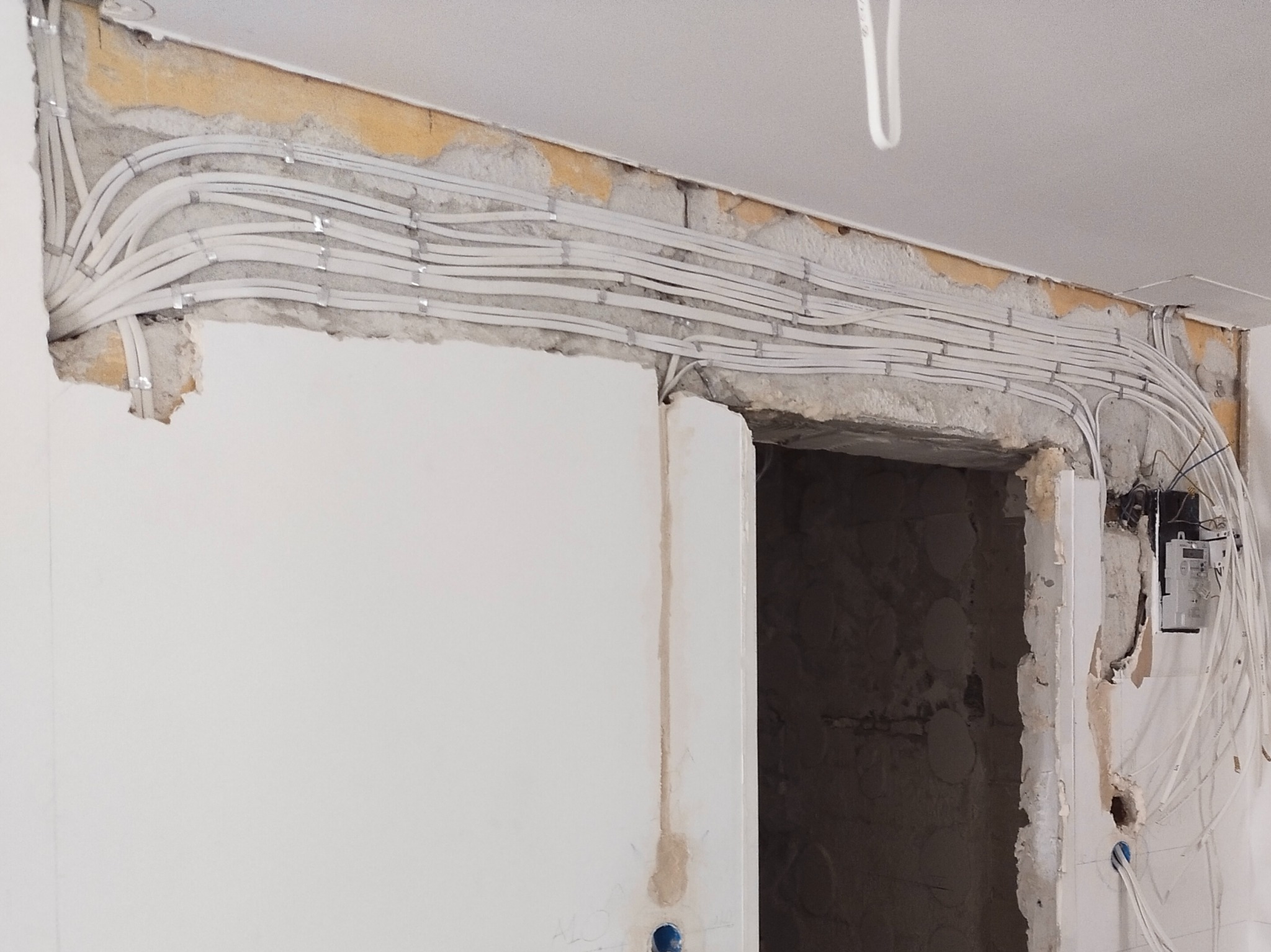 Białe kable elektryczne ułożone równolegle i przymocowane do surowej ściany nad otworem drzwiowym, widoczny licznik elektryczny i wystające przewody.