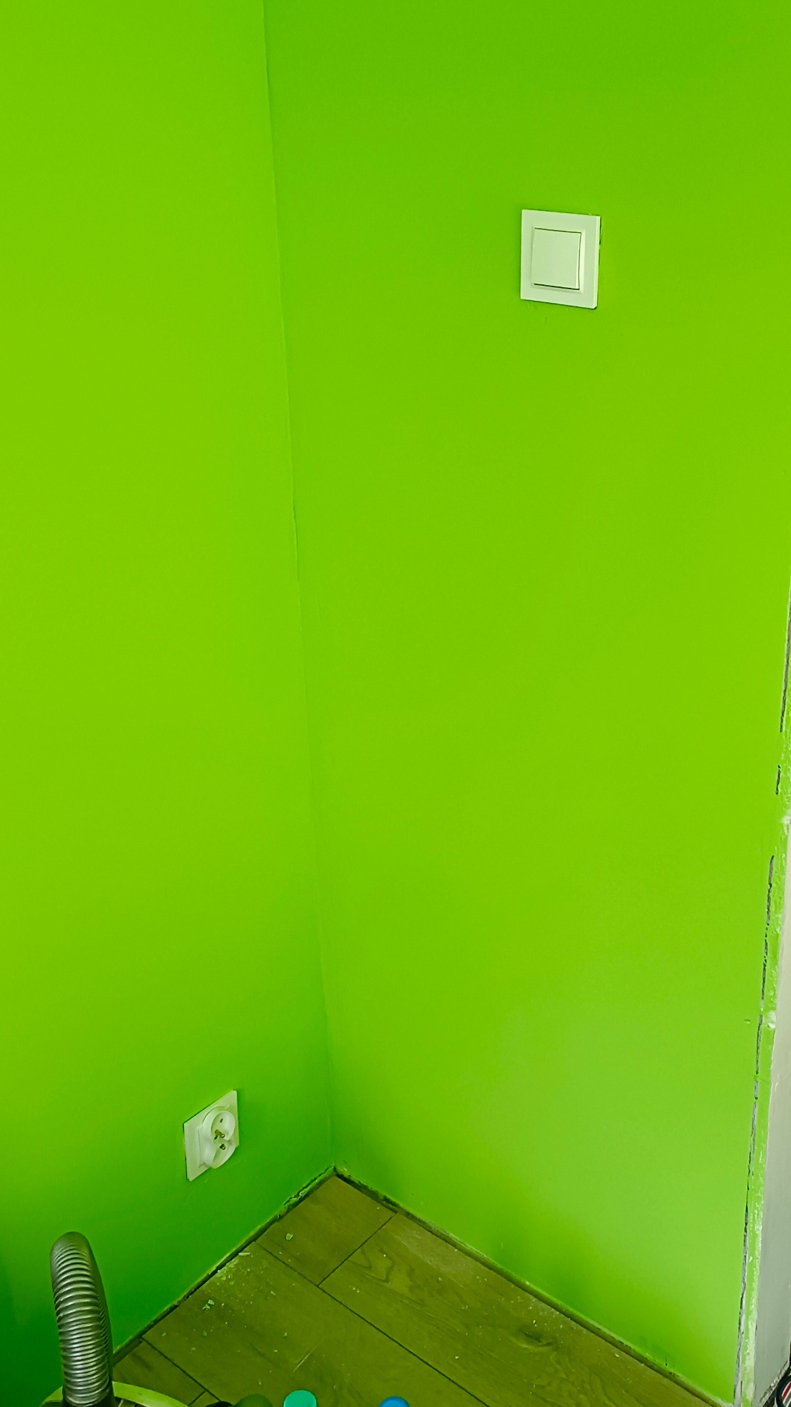 Świeżo pomalowany na jaskrawy zielony kolor róg pokoju z gniazdkiem i włącznikiem. Widoczne ślady farby na podłodze i listwie przypodłogowej.