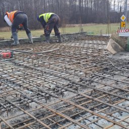 BRM Budownictwo Radosław Mancewicz - Zbrojenie płyty fundamentowej z widocznymi pracownikami w odblaskowych kamizelkach i wibratorem do betonu. Widok na teren budowy z drzewami w tle.