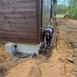 BRM Budownictwo Radosław Mancewicz - Pracownik w trakcie prac ziemnych przy drewnianym domu. Widoczny wykop, narzędzia i fragment fundamentu z folią kubełkową. Prace przygotowawcze do izolacji fundamentów.