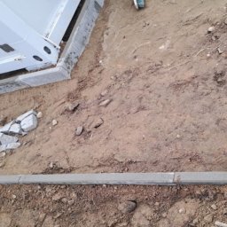 BRM Budownictwo Radosław Mancewicz - Etap układania kostki granitowej: obrzeża ułożone, podłoże przygotowane, kostka częściowo ułożona przy budynku. Widoczne narzędzia i materiały budowlane.